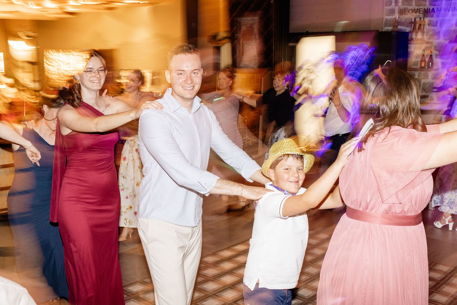 Wedding guests including a child in a hat dance together joyfully on a colorful reception dance floor.