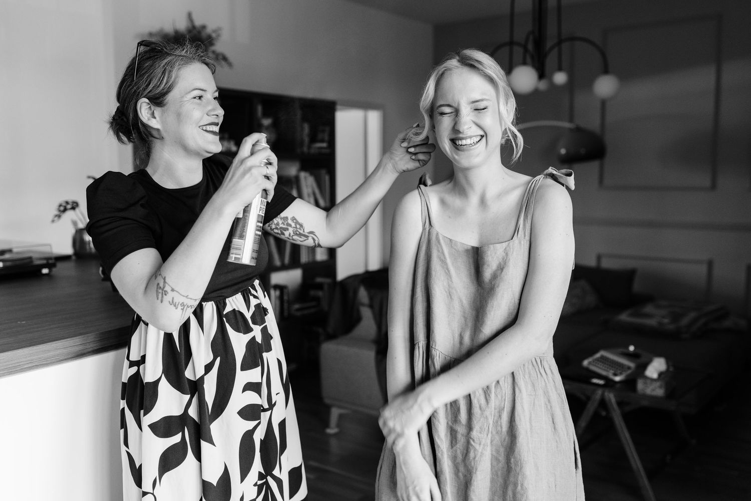 Black and white photo of a makeup artist styling a laughing blonde woman in a floral skirt indoors.