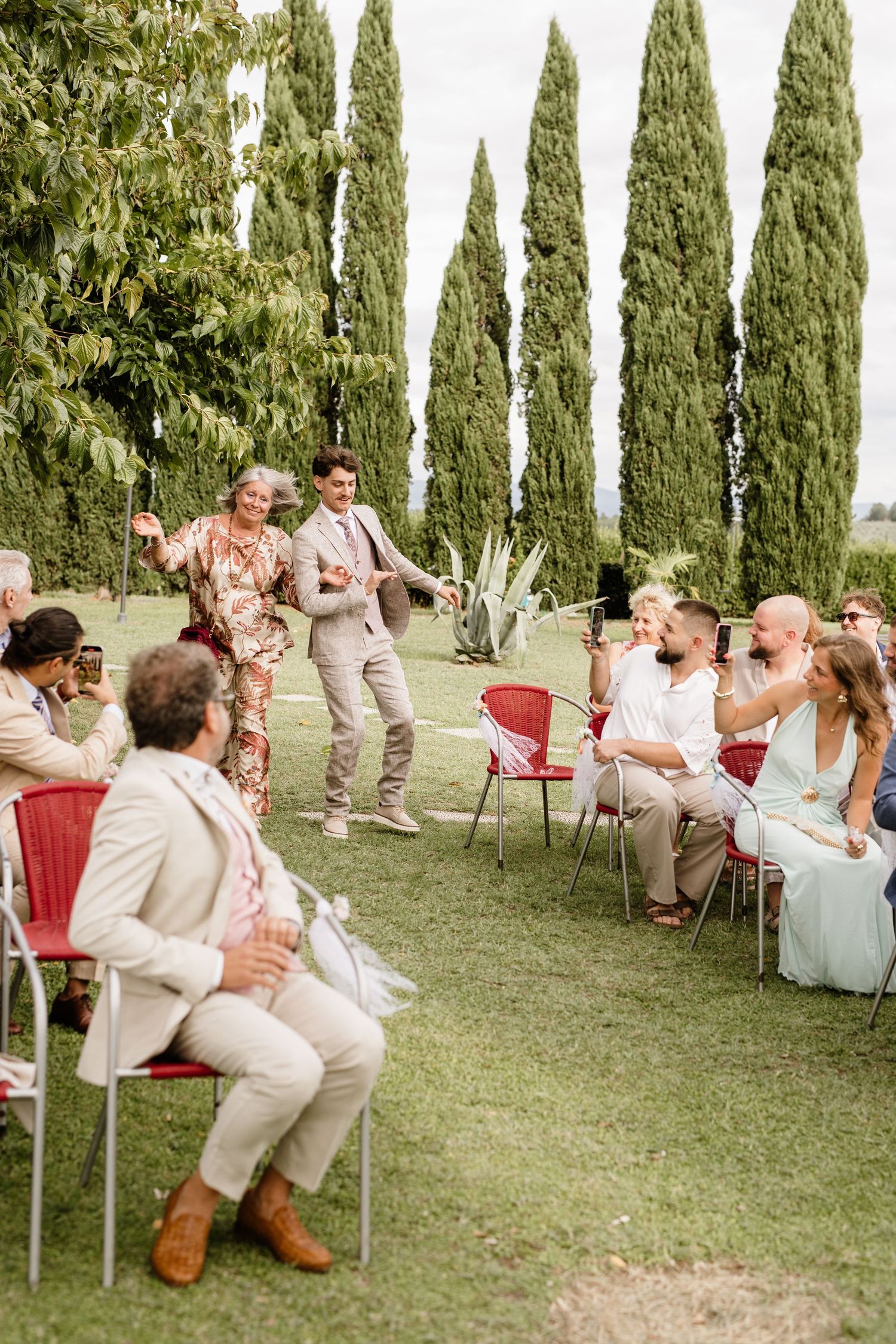 Guests celebrate as two grooms walk down the aisle at an outdoor wedding ceremony with cypress trees.