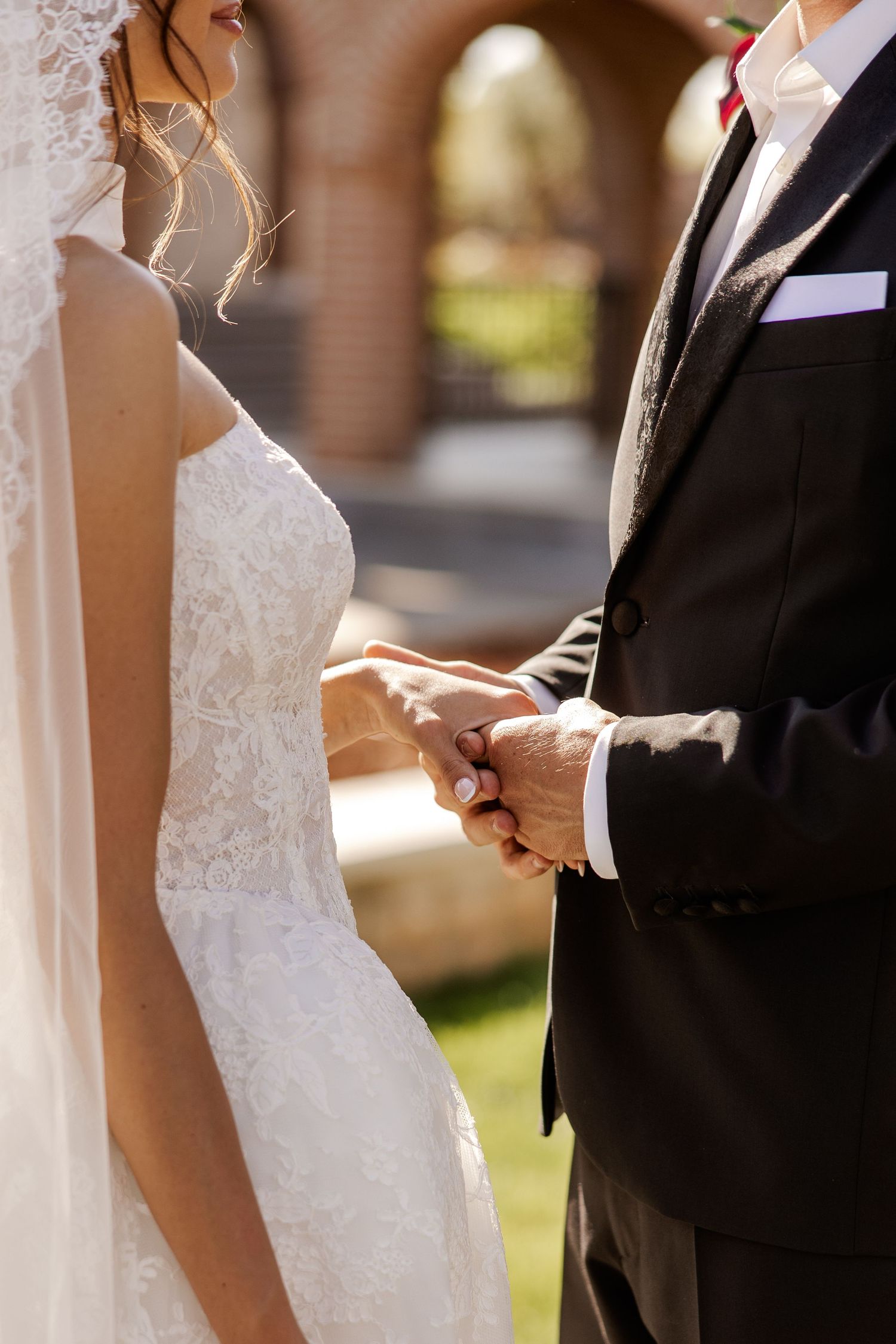 Bride and groom holding hands during outdoor wedding ceremony, wearing white lace gown and black suit.