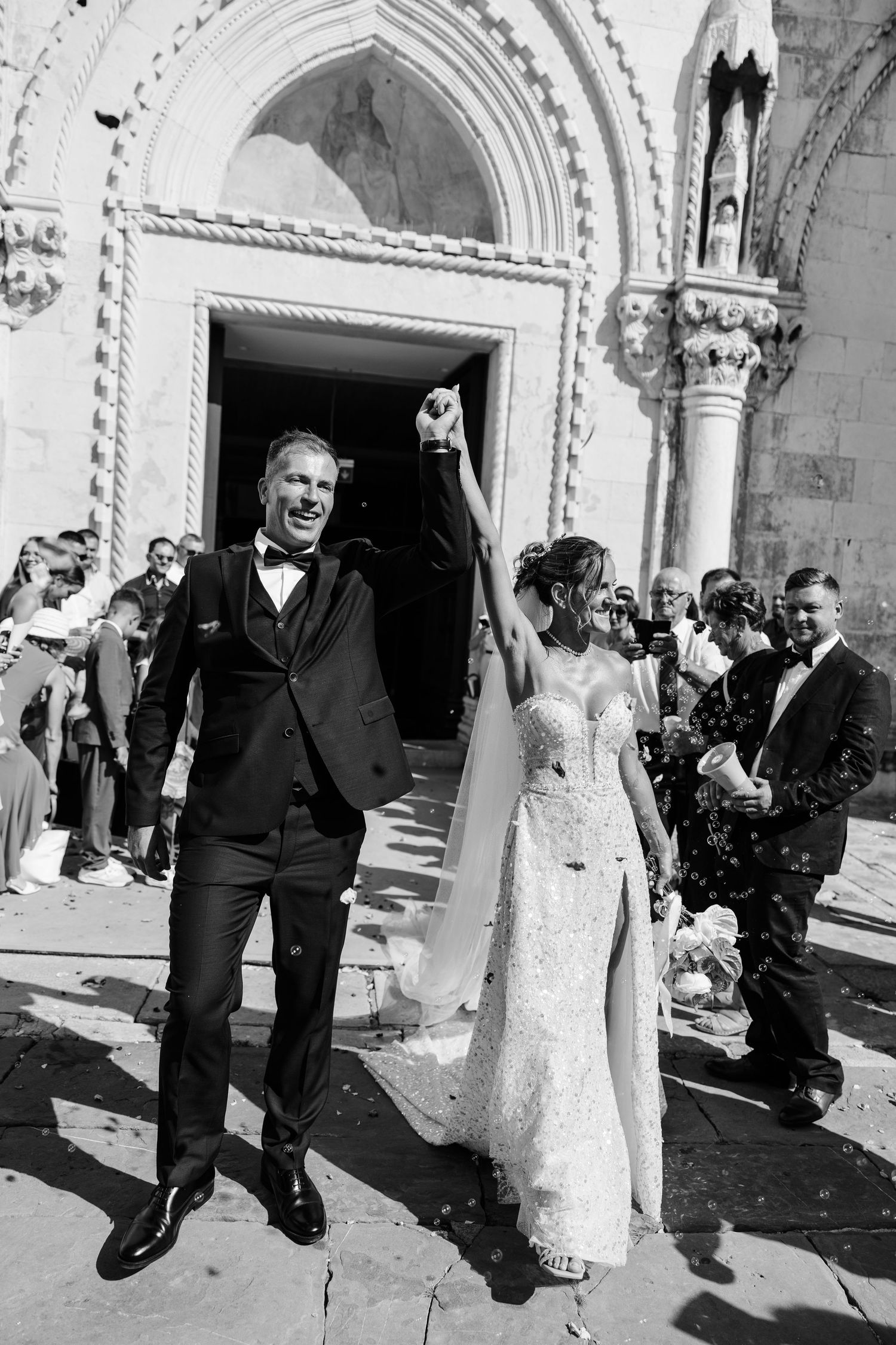 Newlyweds celebrate joyfully outside a historic church, groom in tuxedo raises bride's hand in black and white photo.