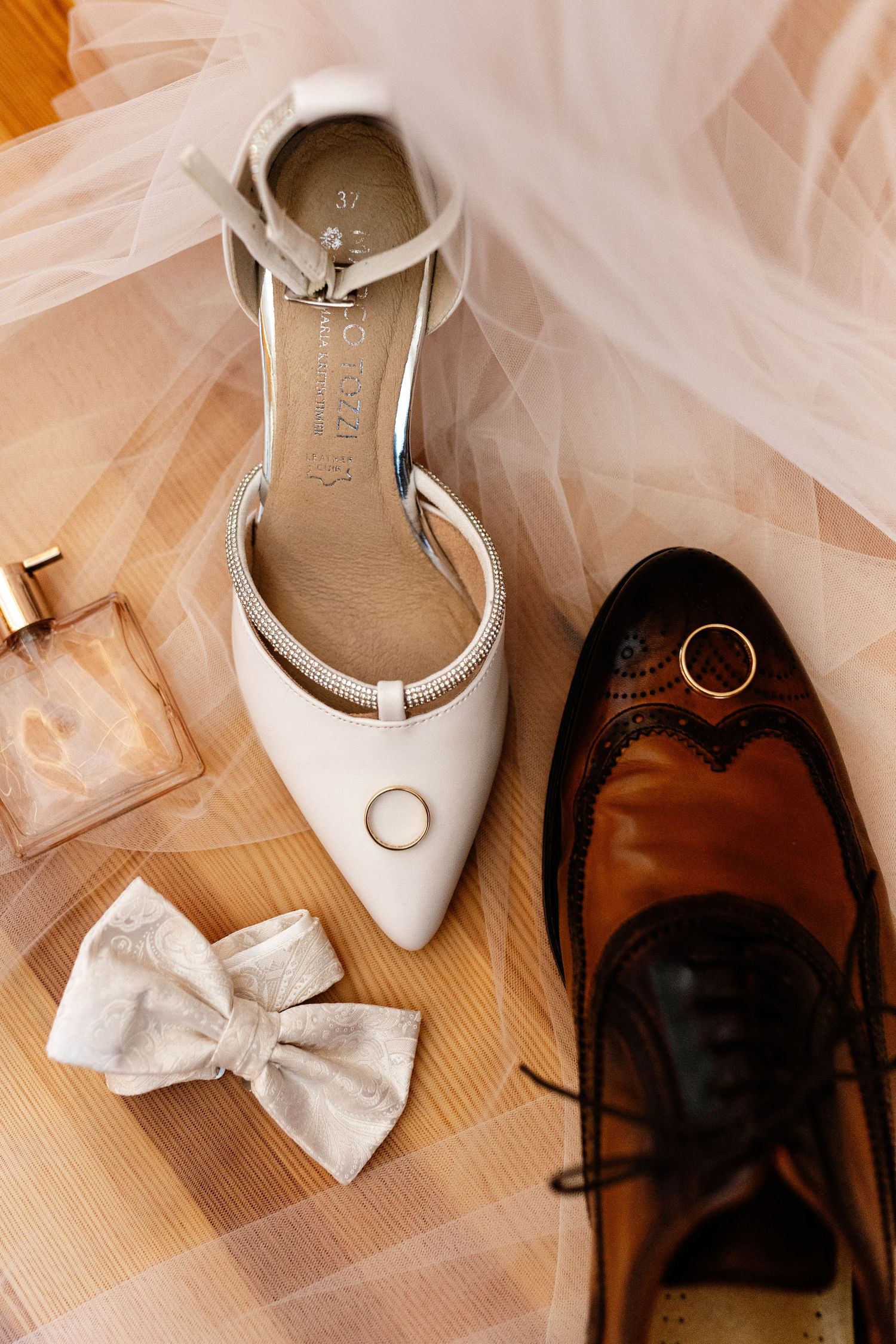 Wedding flat lay with white bridal heels, brown groom shoes, rings, bow tie, and perfume on wood floor.