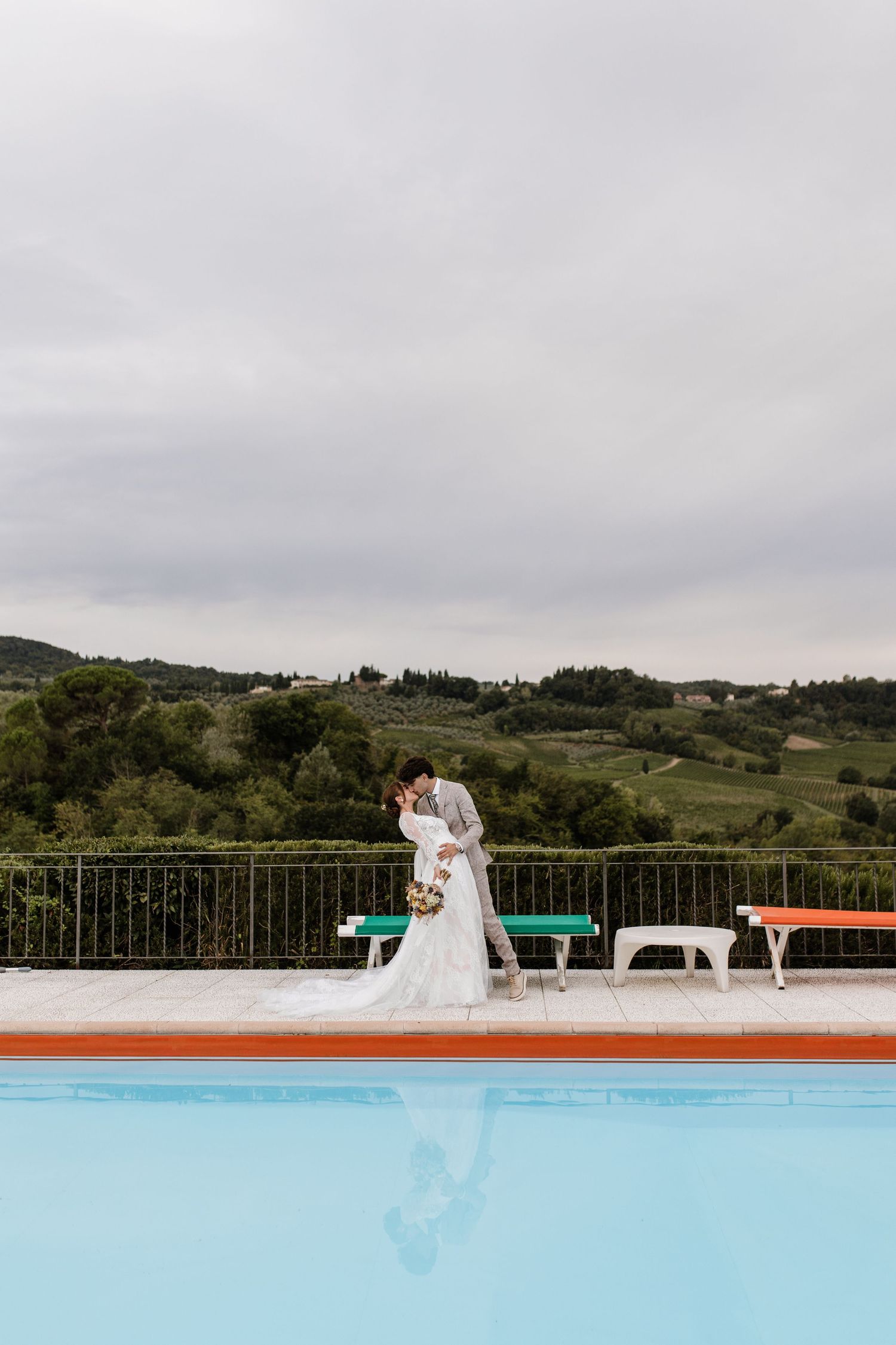 Bride and groom share a romantic kiss beside a pool with rolling Tuscan hills in the background.