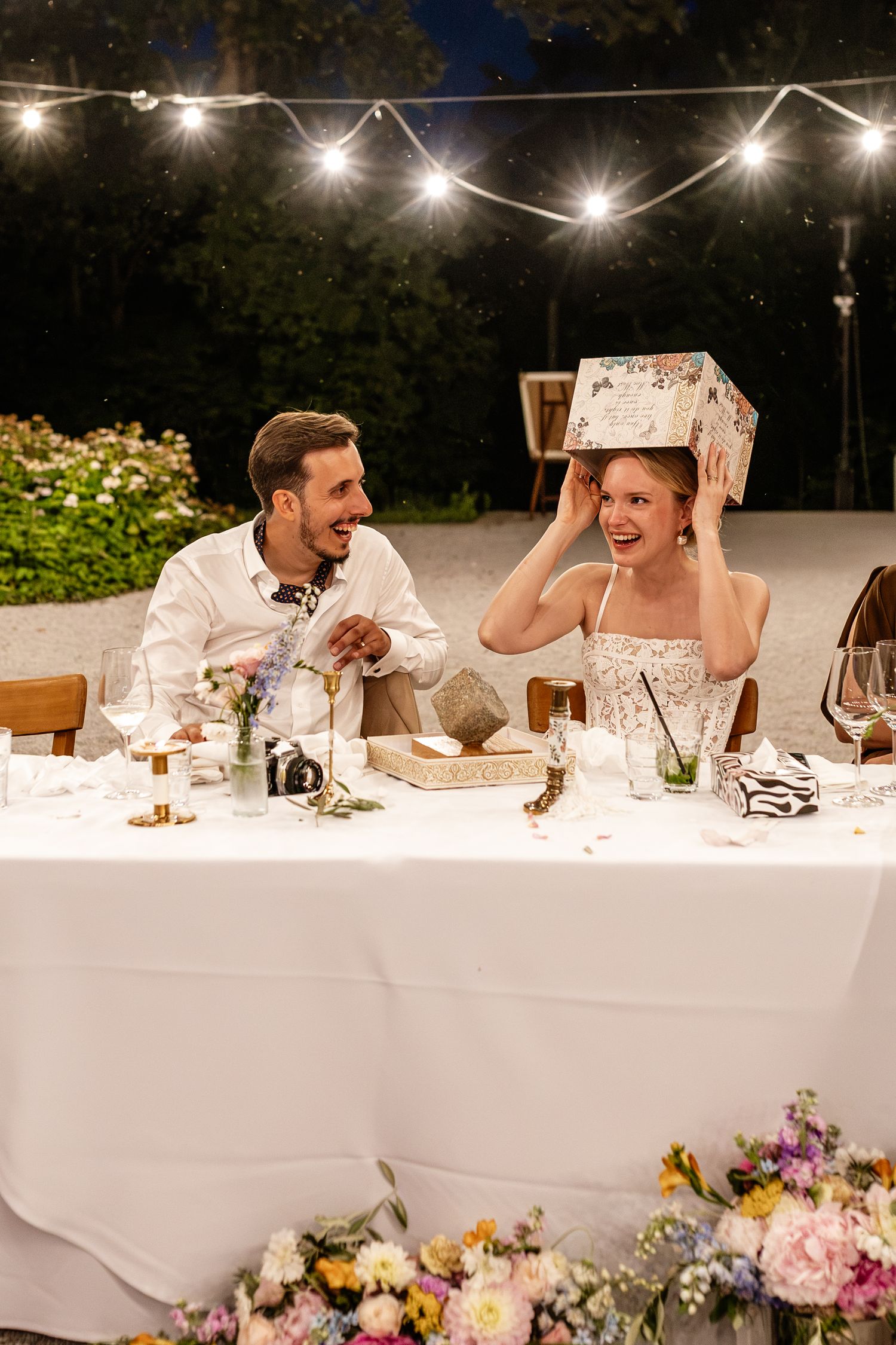 Bride and groom laughing at reception table under string lights as bride playfully holds gift box over her head.