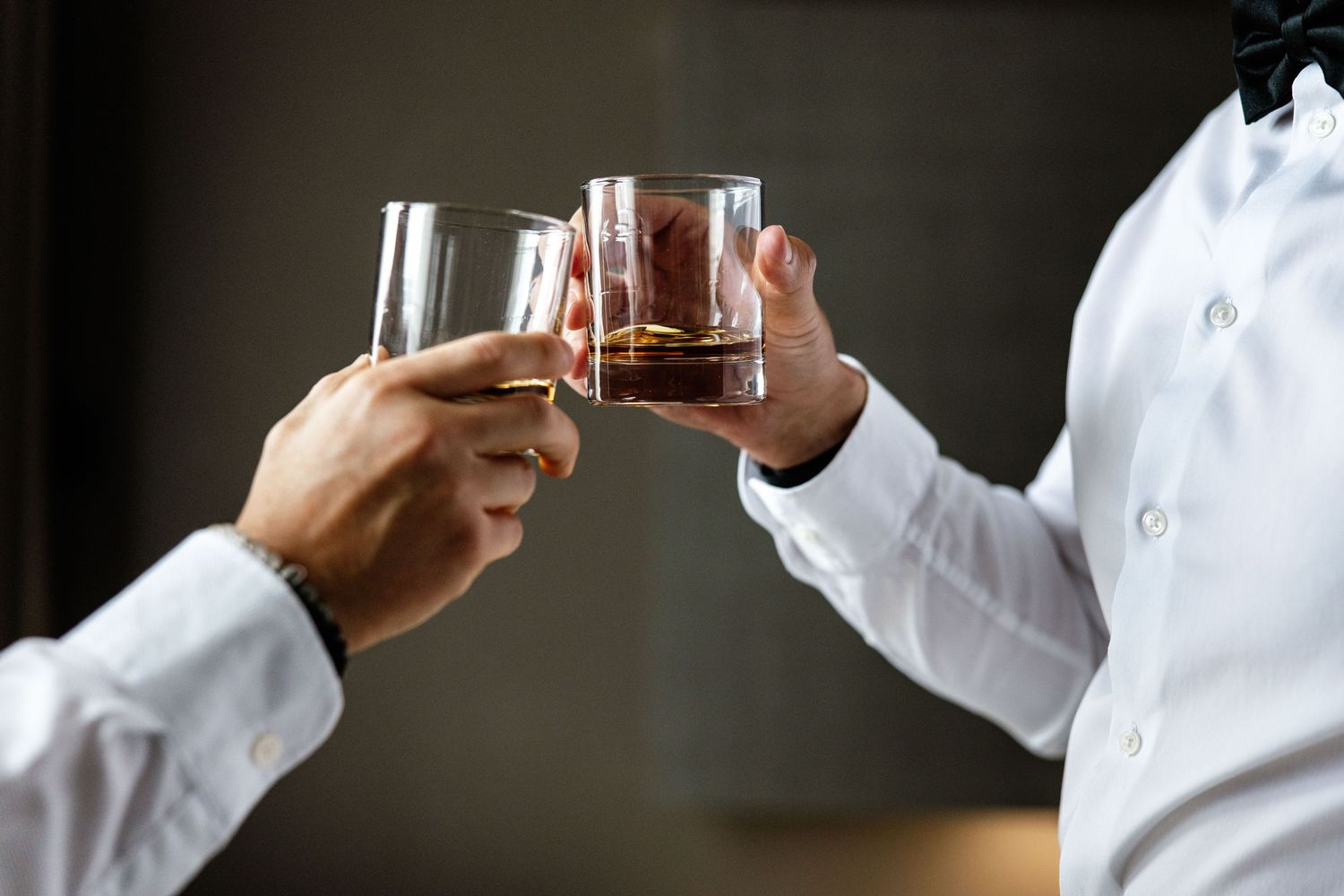 Two men in white dress shirts toasting with glasses of whiskey or bourbon in a celebratory cheers.