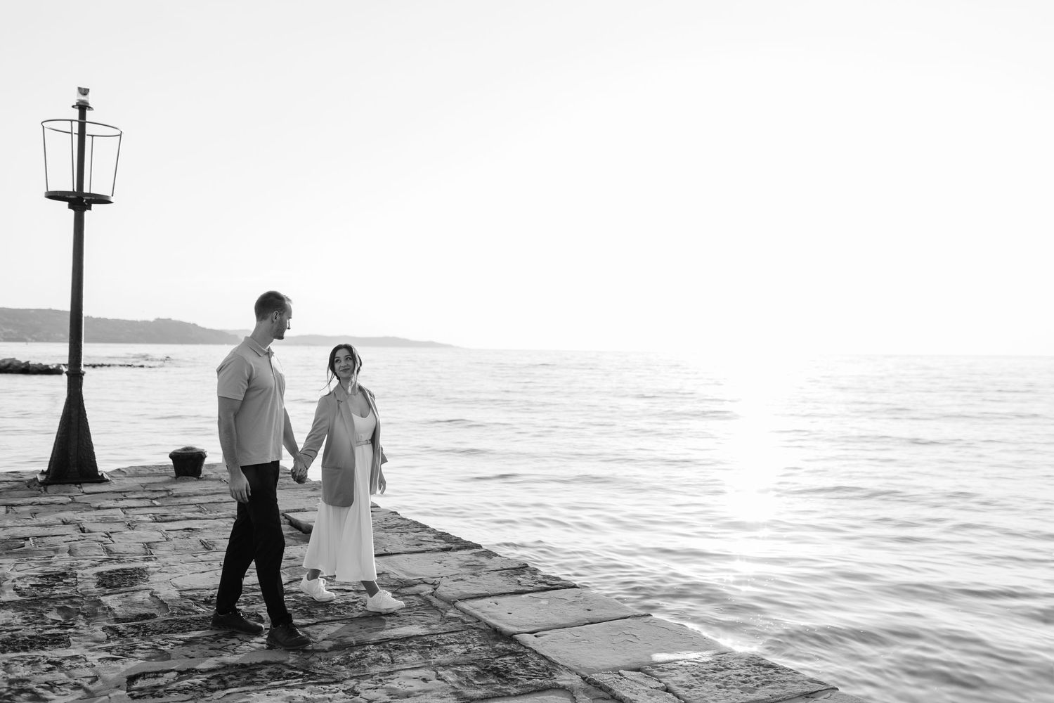Black and white photo of a couple holding hands walking along a sunlit seaside pier.