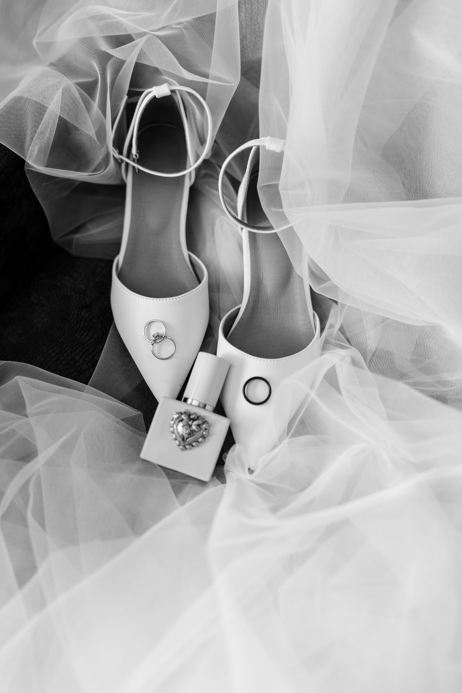 Black and white photo of bridal heels, wedding rings, and perfume bottle arranged on a flowing white veil.