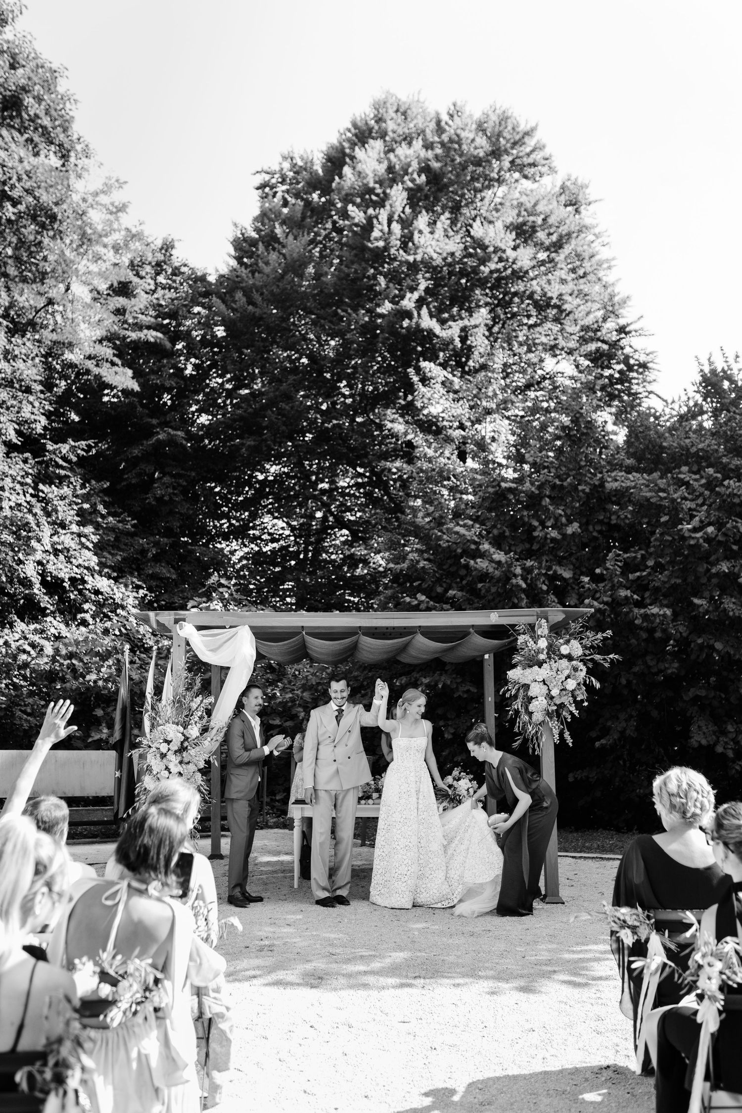 Black and white photo of bride and groom at outdoor wedding ceremony beneath floral arch surrounded by guests.