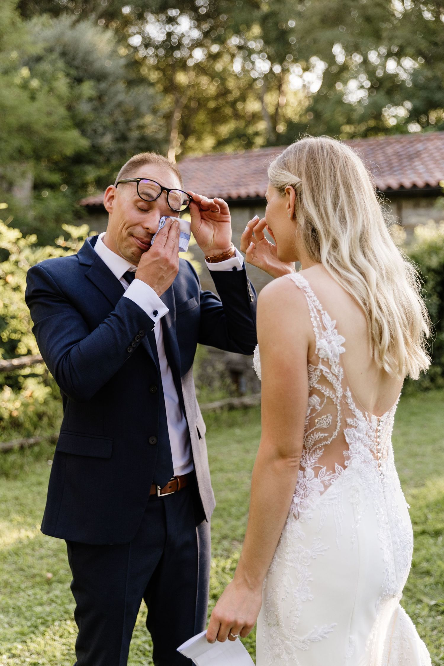 Emotional groom lifting glasses to wipe tears during first look with blonde bride outdoors.