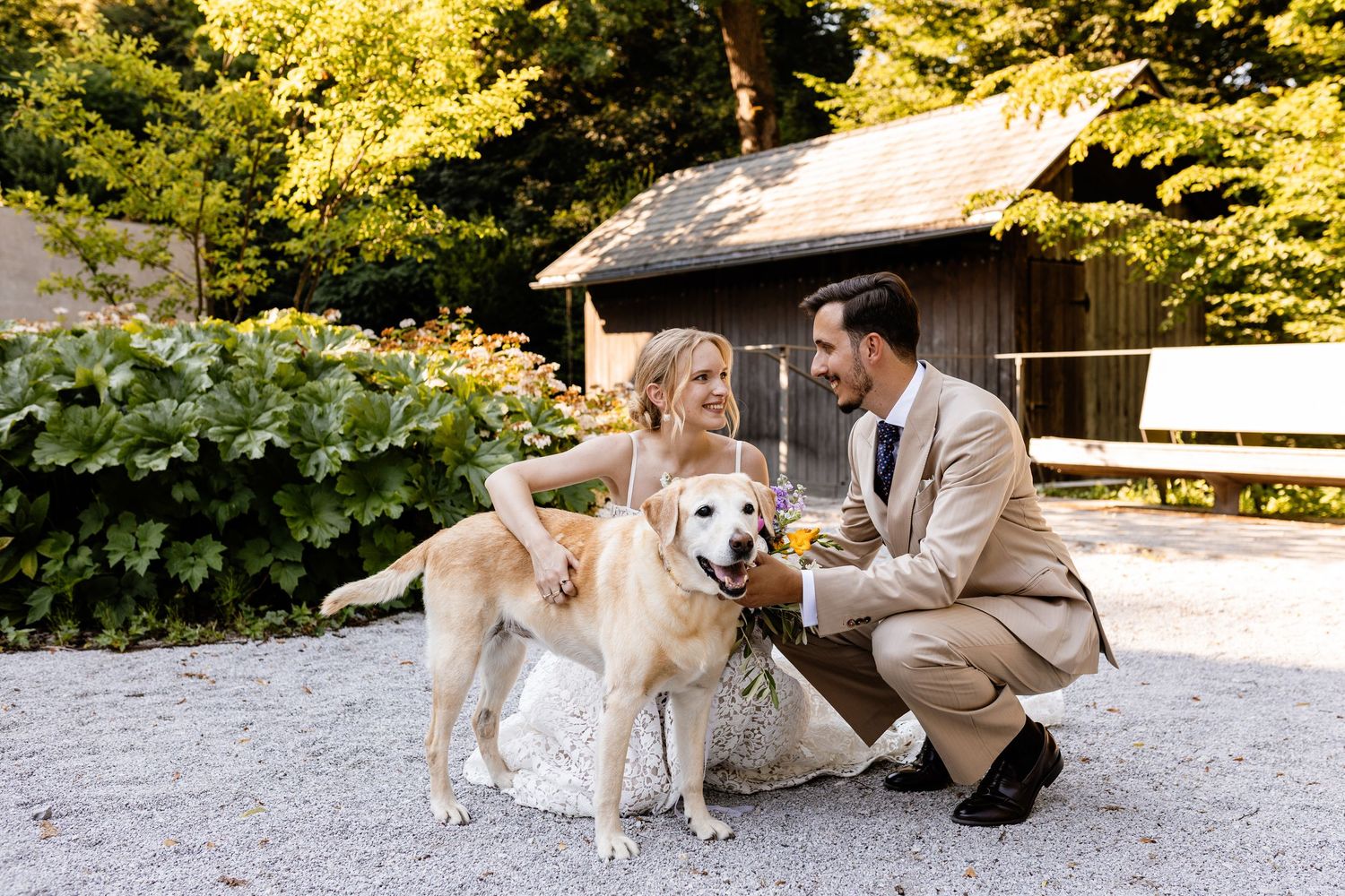 Happy newlyweds crouch beside their golden retriever dog outdoors, holding champagne glasses at rustic wedding.