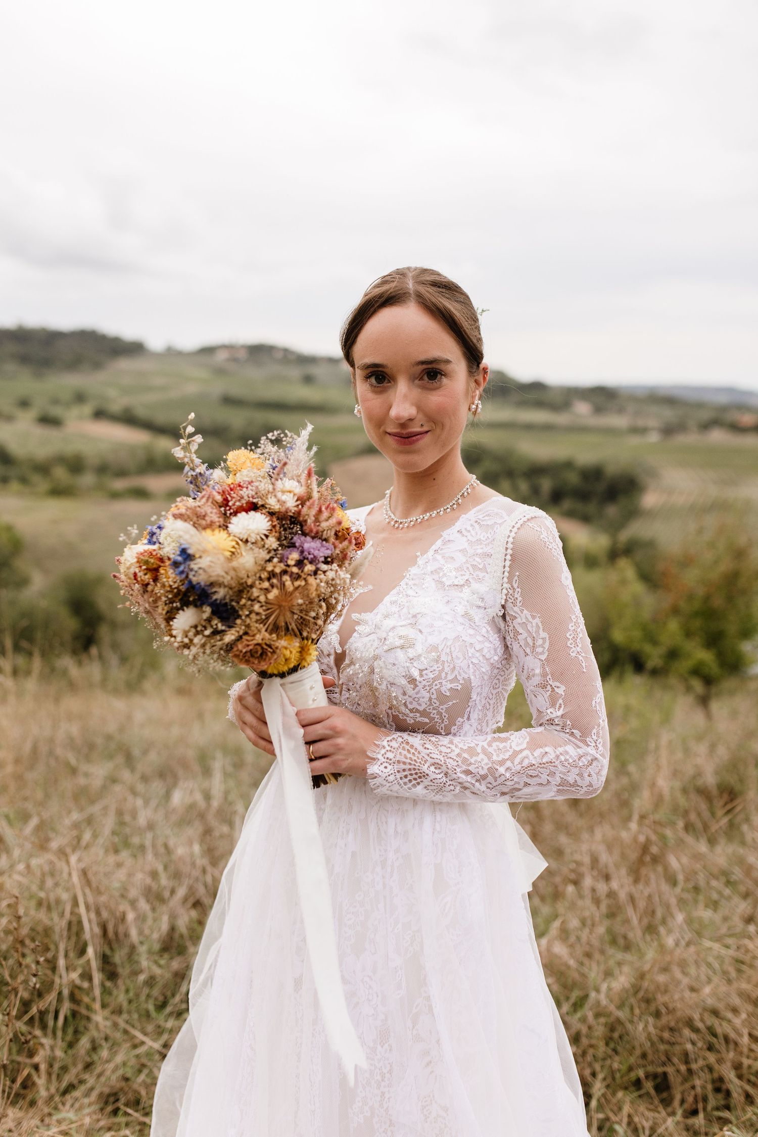 Bride in lace wedding gown holds colorful dried flower bouquet in a scenic countryside field.