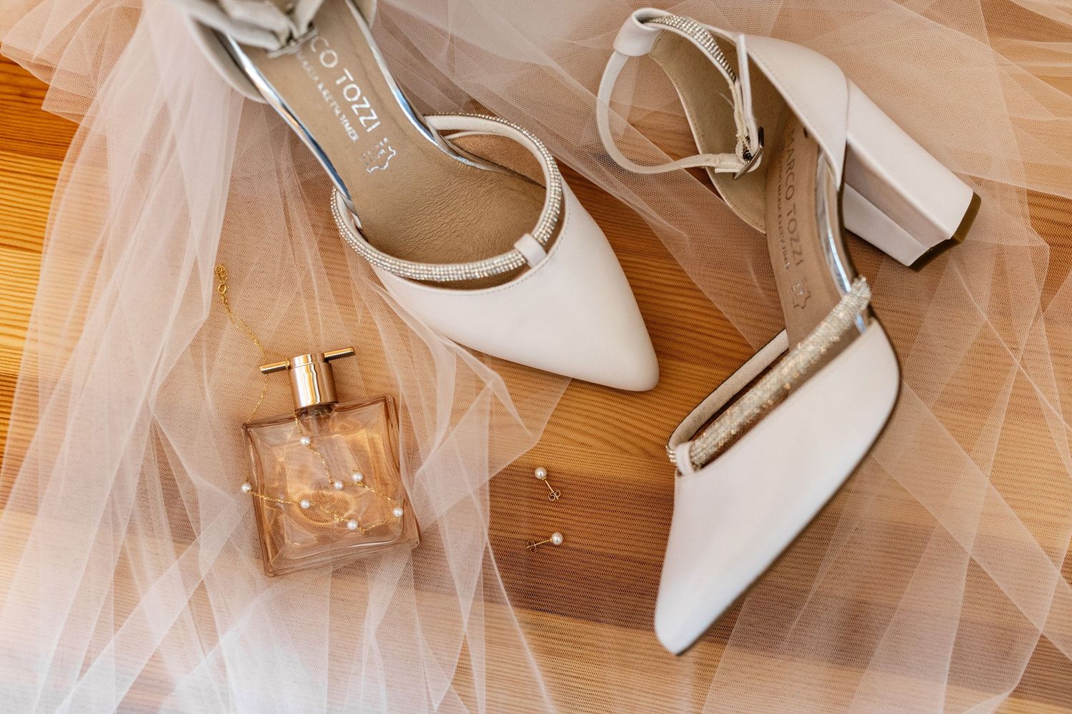 White block heel bridal shoes with ankle straps beside a gold perfume bottle on sheer tulle fabric.