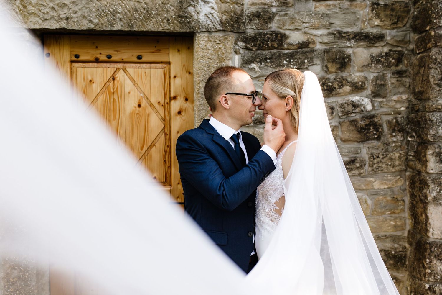 A groom in a navy suit tenderly holds his bride's face near a rustic stone wall and wooden door.