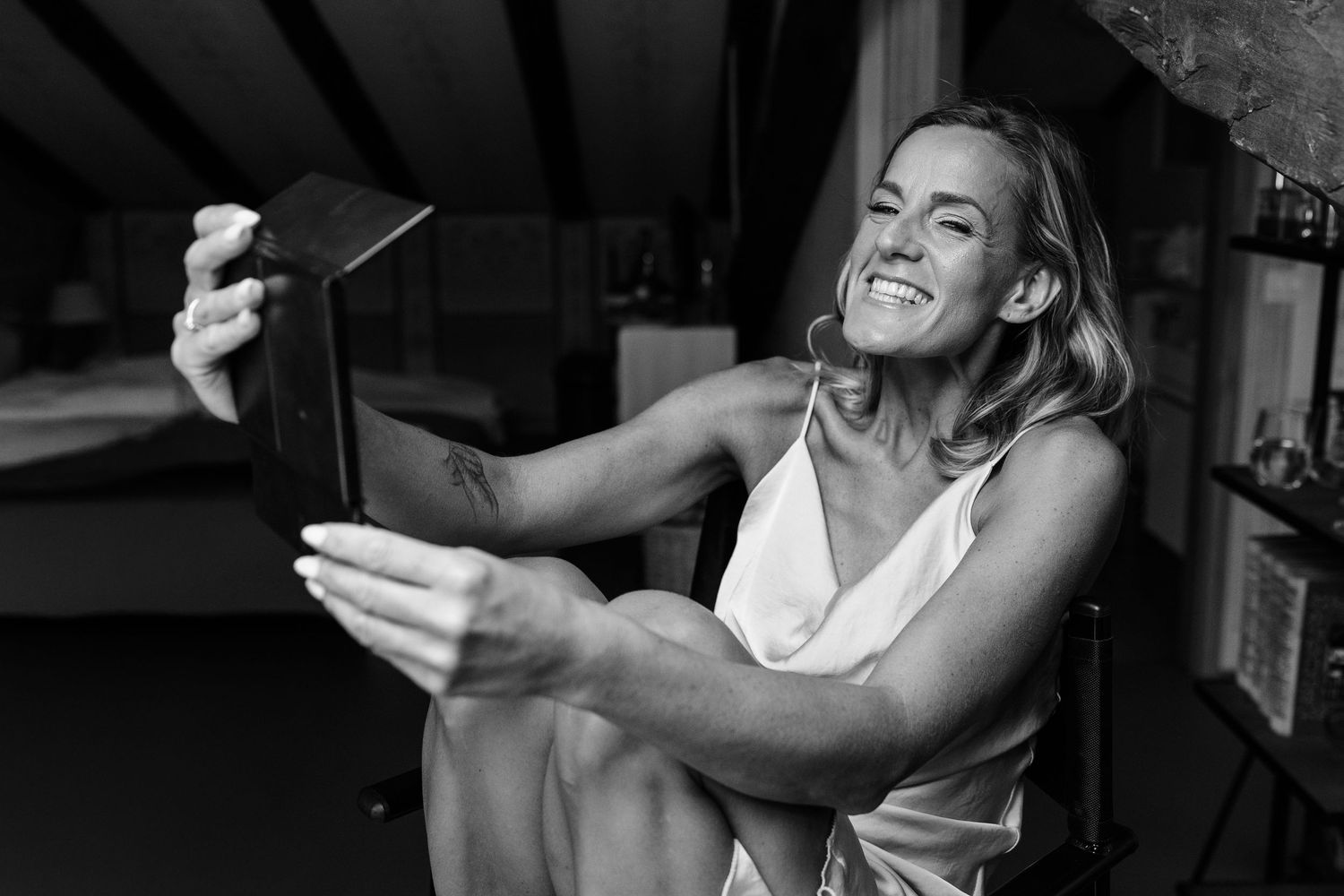 Black and white photo of a smiling blonde woman taking a selfie with a tablet while sitting indoors.