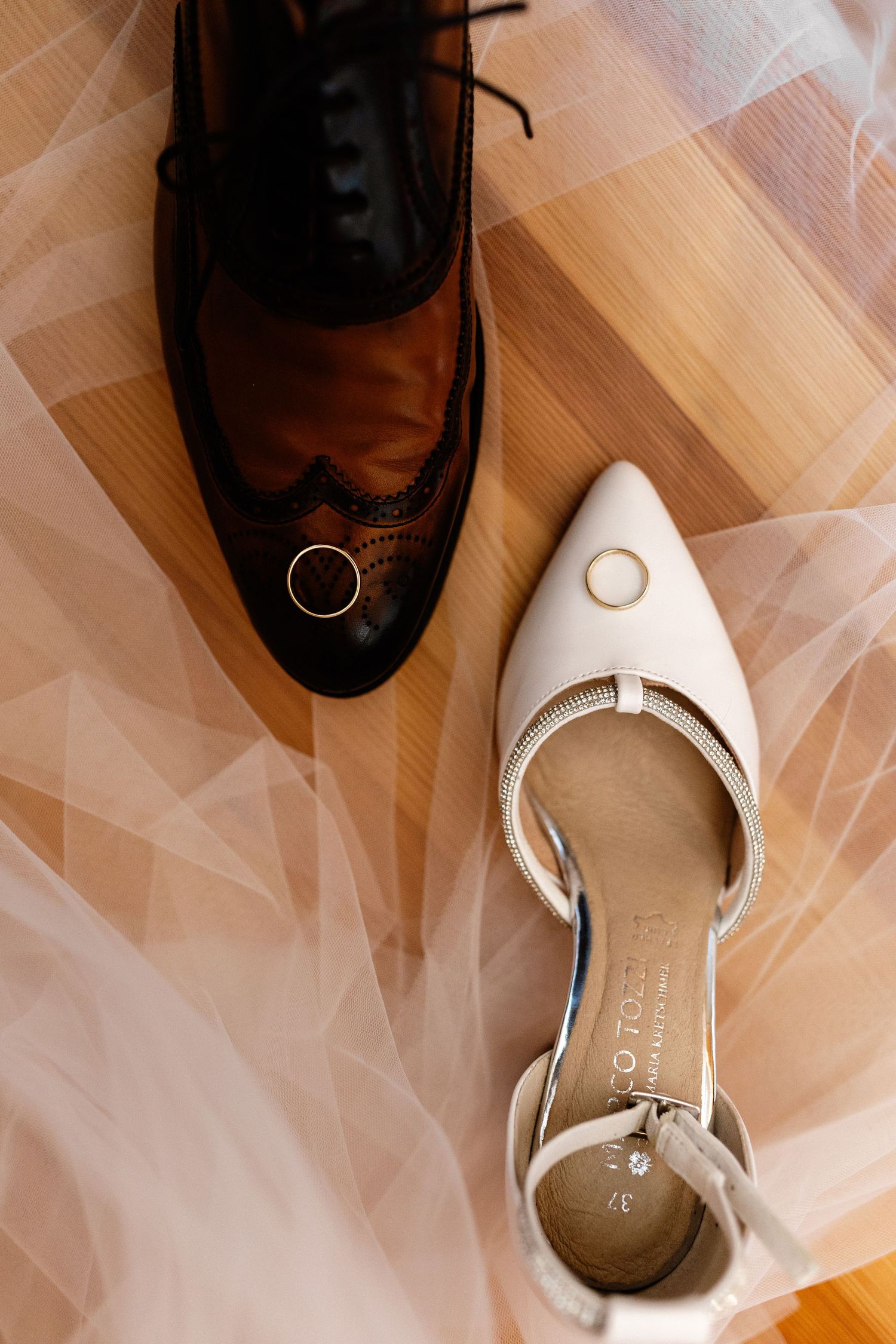Wedding rings resting on brown brogue and white pointed-toe bridal heels on wooden floor with tulle veil.