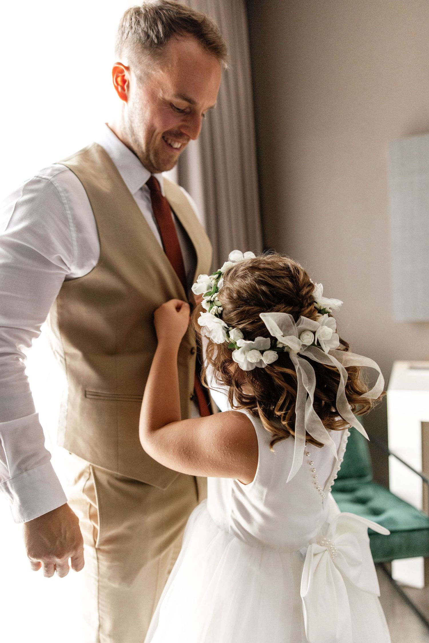 Groom in beige vest smiling as flower girl with white floral crown helps him prepare.