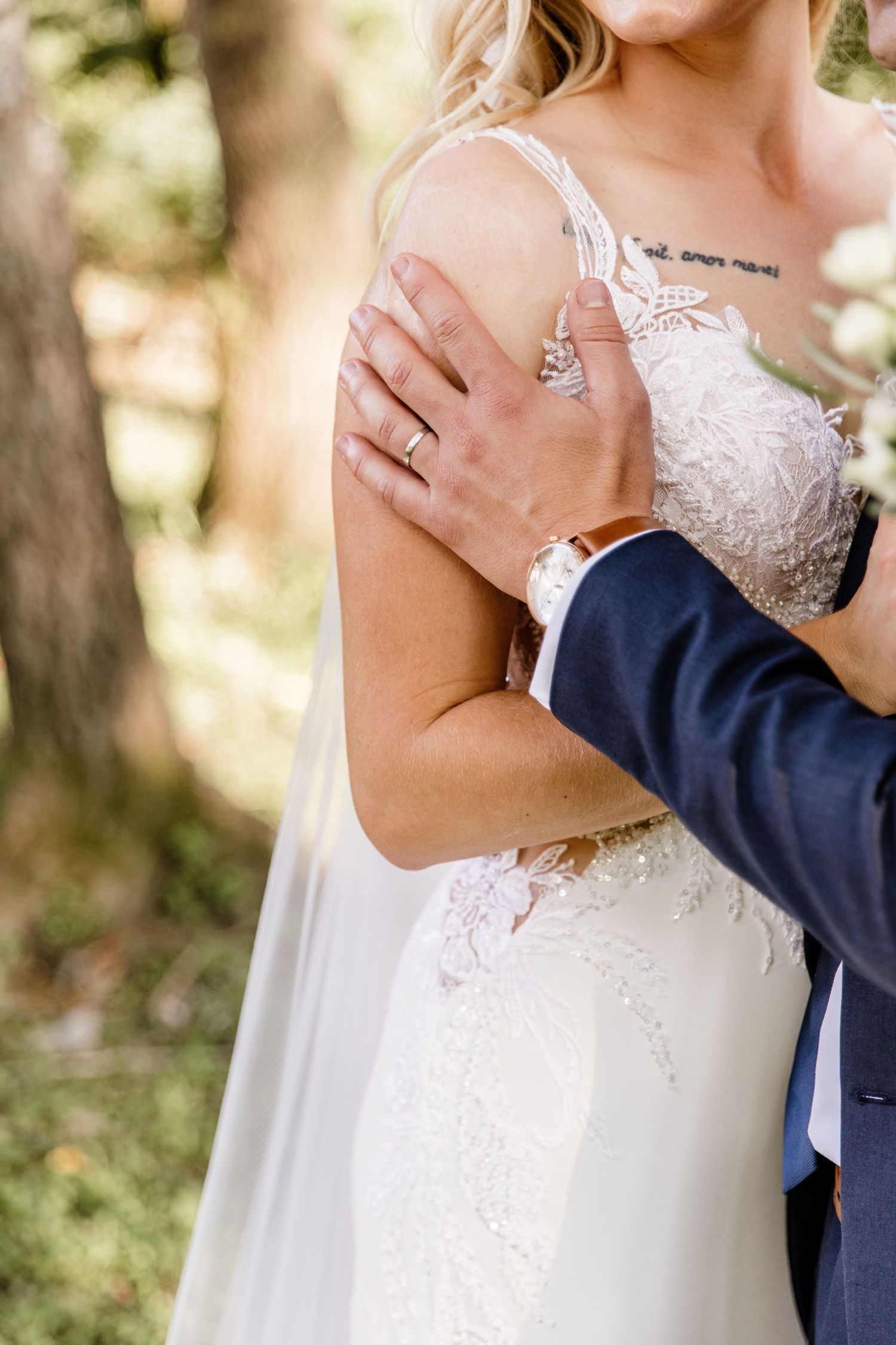 Groom in navy suit embracing bride in lace wedding dress, showing wedding rings and floral bouquet outdoors.