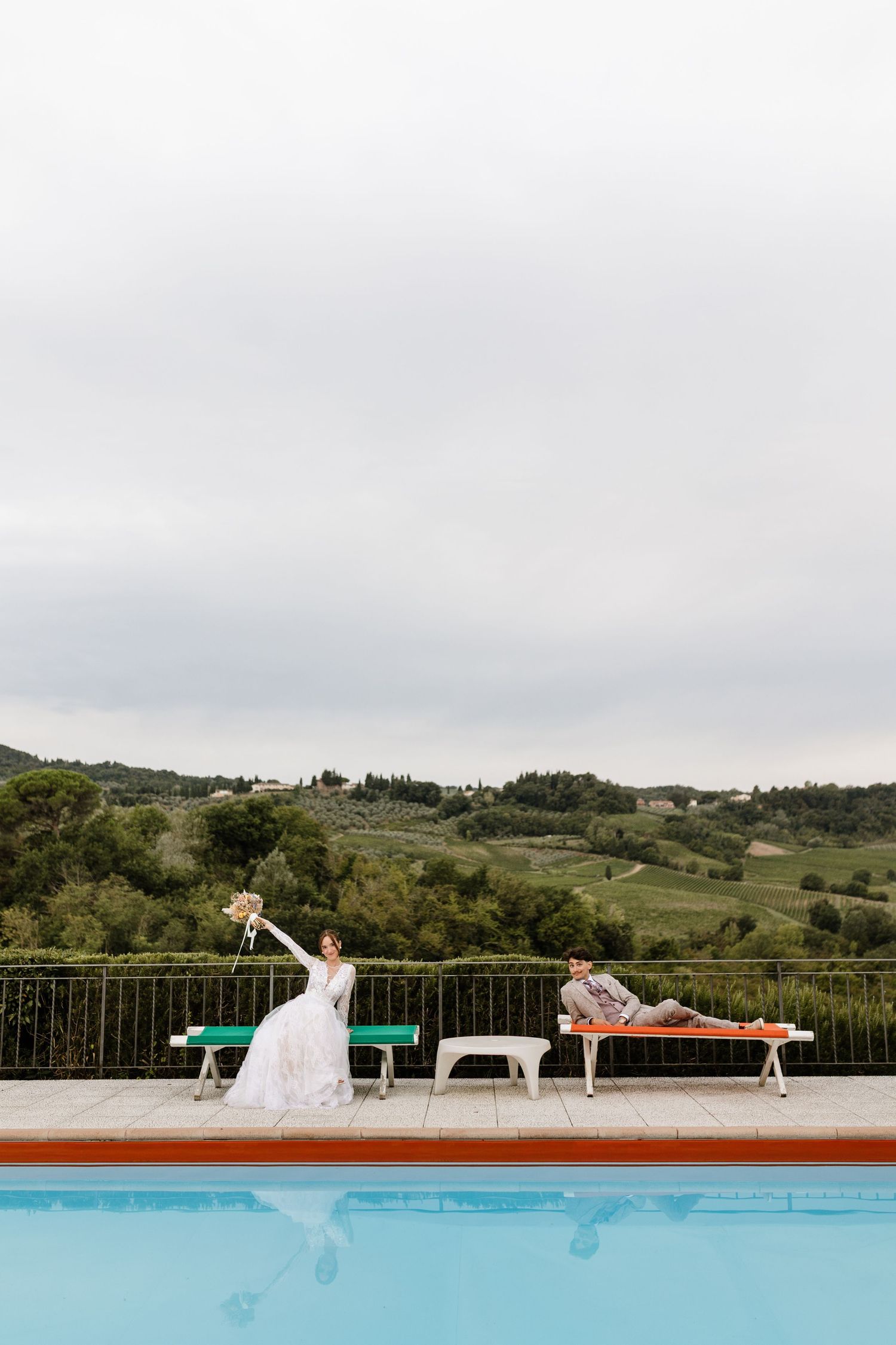 Bride and groom relaxing by a pool with rolling vineyard hills in the background.