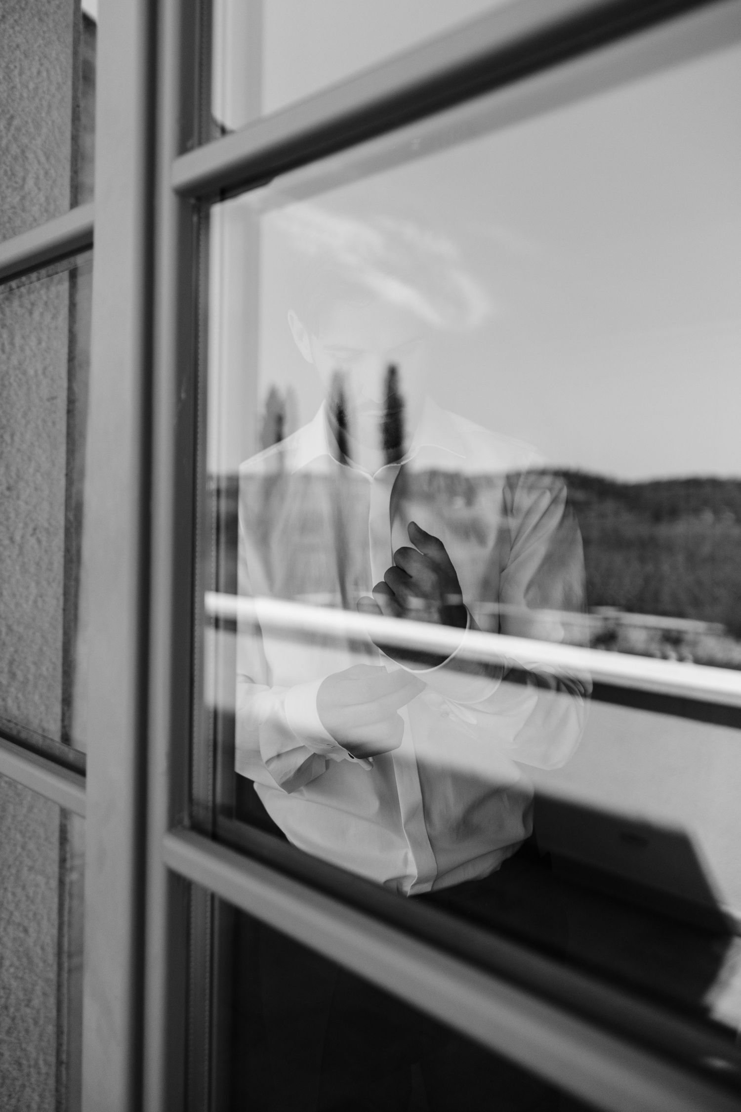 Black and white photo of a groom adjusting his tie reflected in a window.