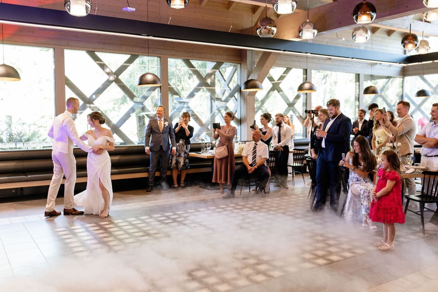 Elegant wedding reception with guests watching couple share first dance on checkered floor in bright venue.