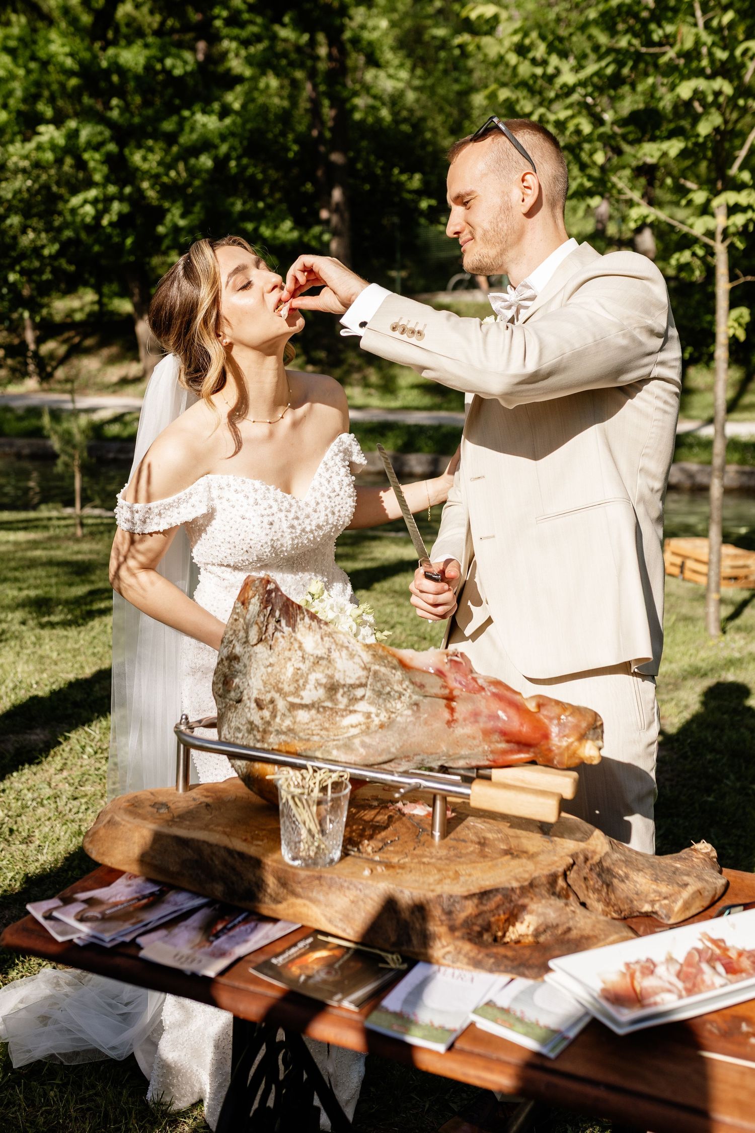 Groom in beige suit feeds bride a bite of food at outdoor wedding reception charcuterie display.
