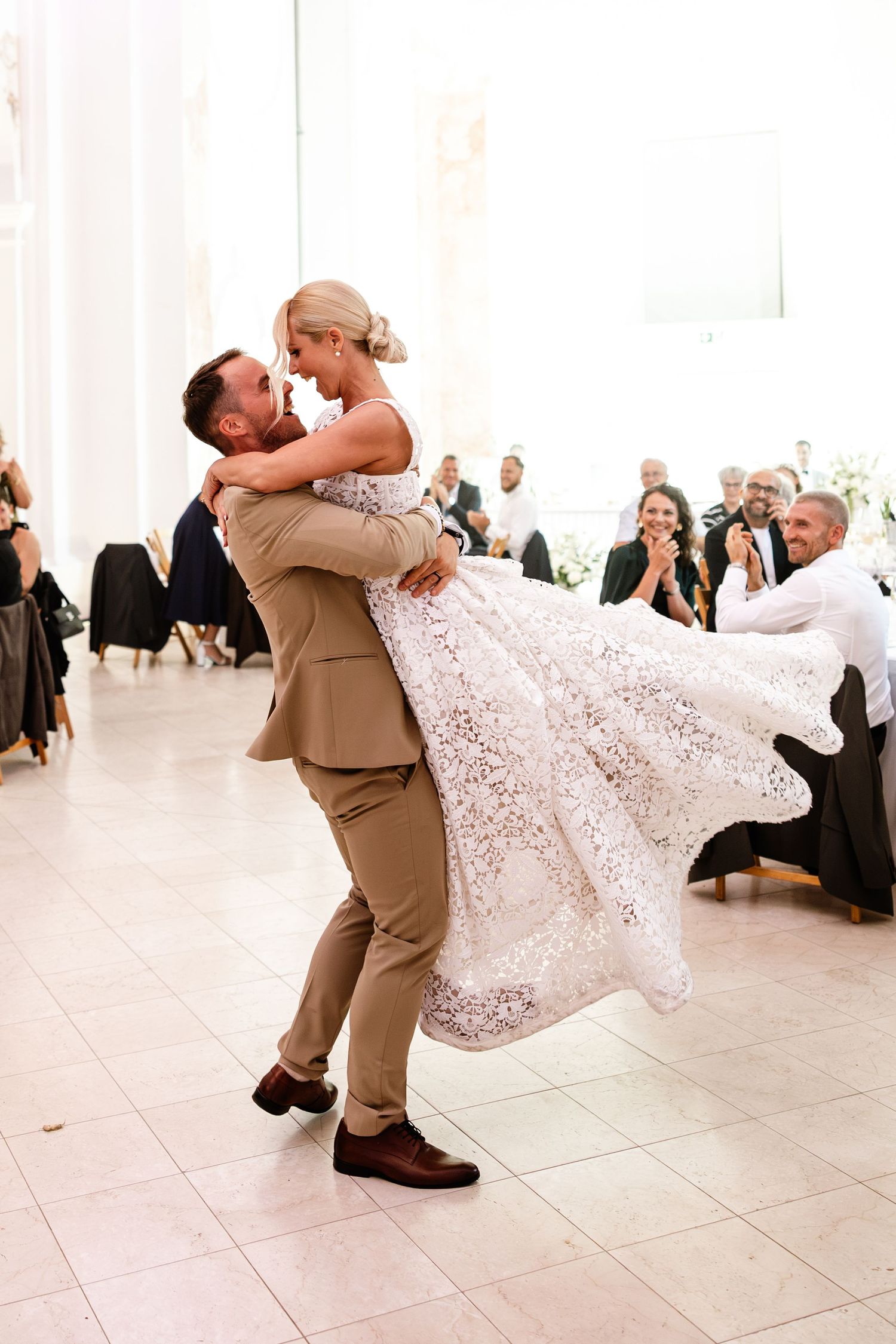 Groom in tan suit lifts bride in lace wedding dress during romantic first dance at wedding reception.