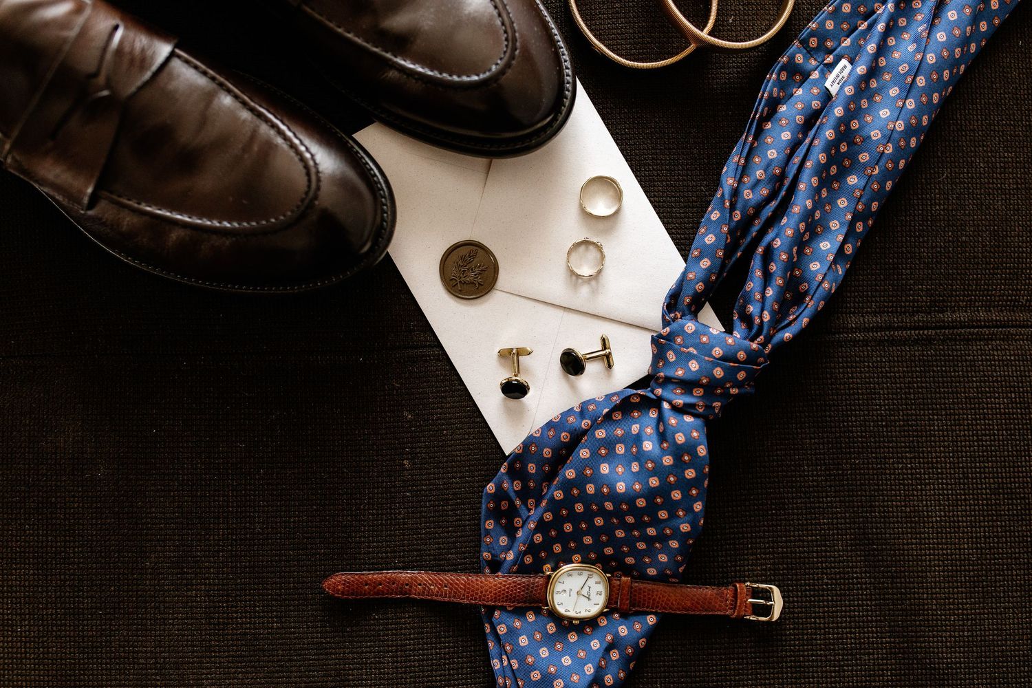 Flat lay of groom wedding accessories including brown leather shoes, blue polka dot tie, watch, cufflinks, and gold rings.