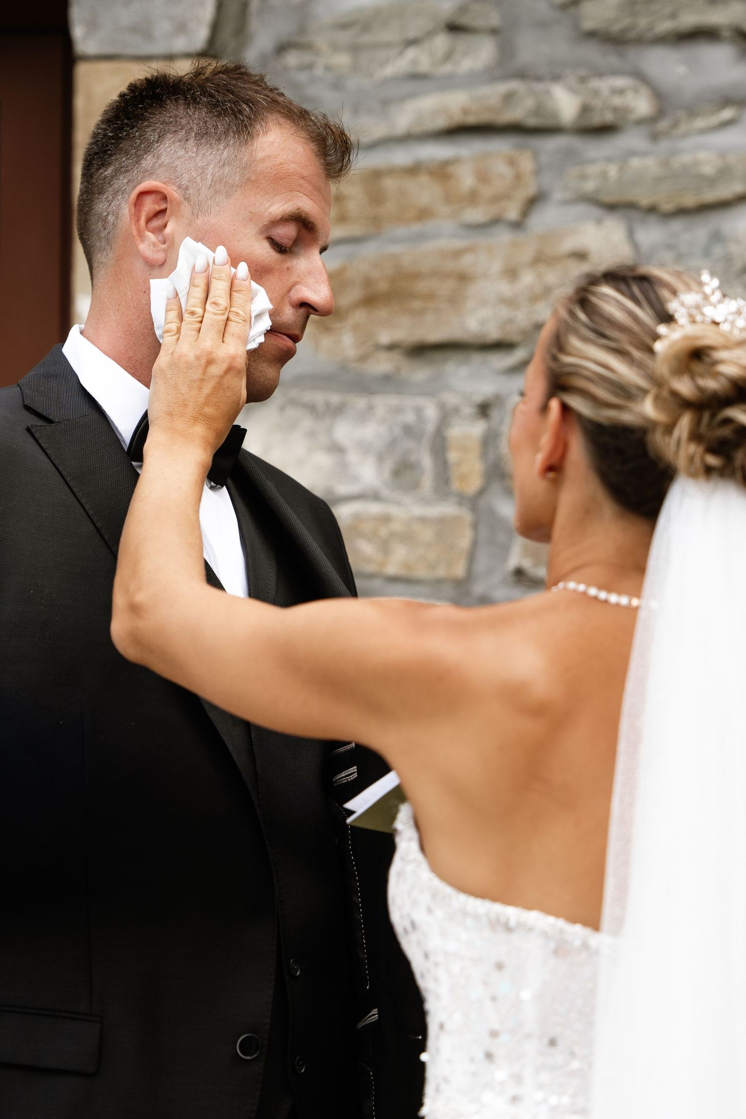 Emotional groom being wiped of tears by bride during outdoor wedding ceremony against stone wall.