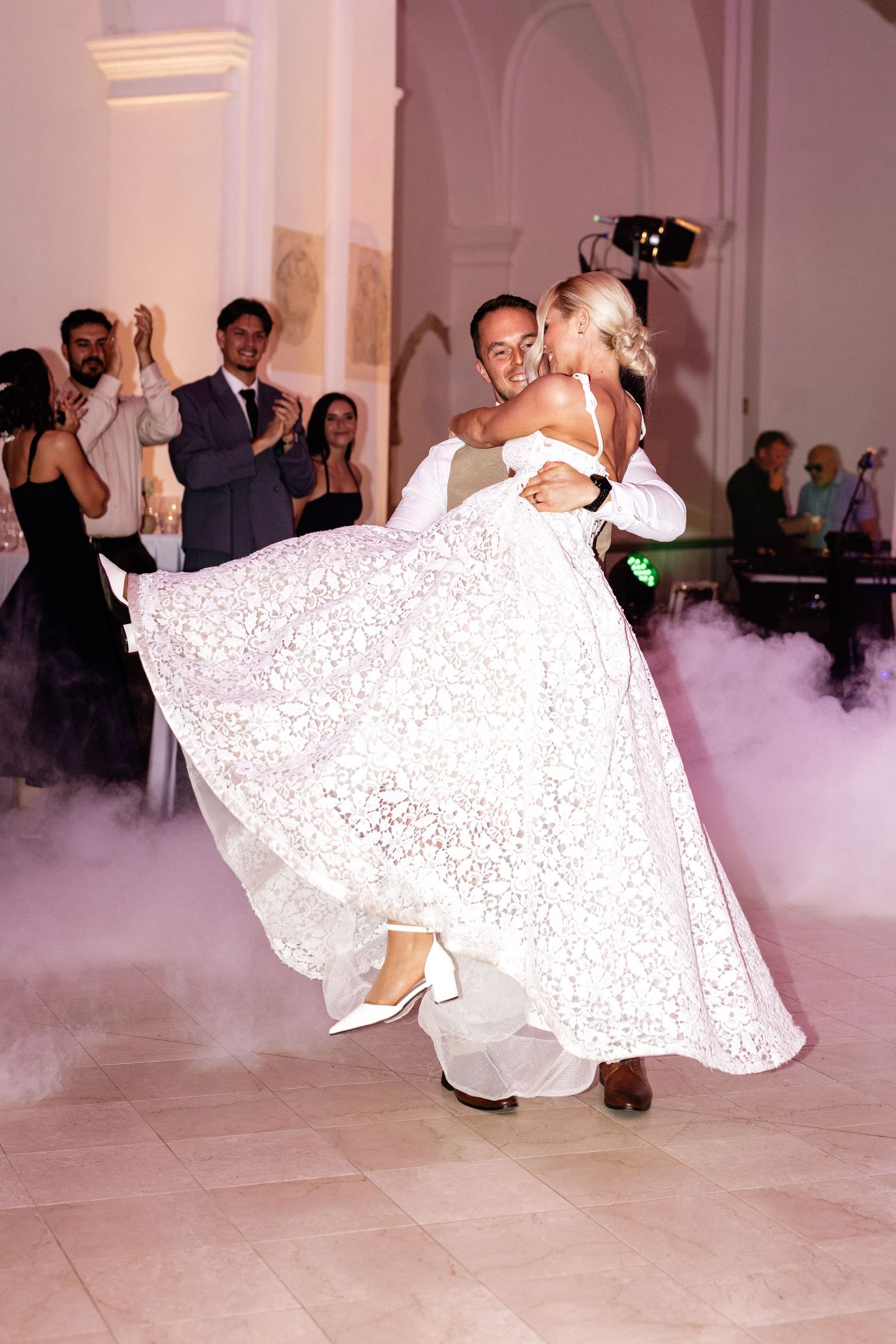 Groom lifting bride in white lace gown during first dance, surrounded by guests in elegant venue with misty floor effects.
