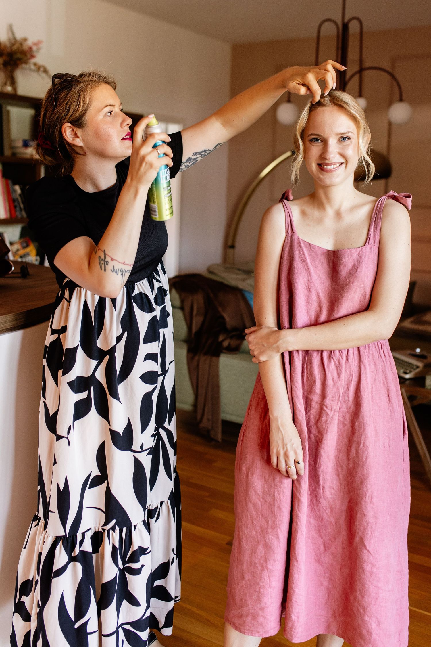 Woman in floral skirt applying hairspray to smiling blonde friend in pink linen dress indoors.