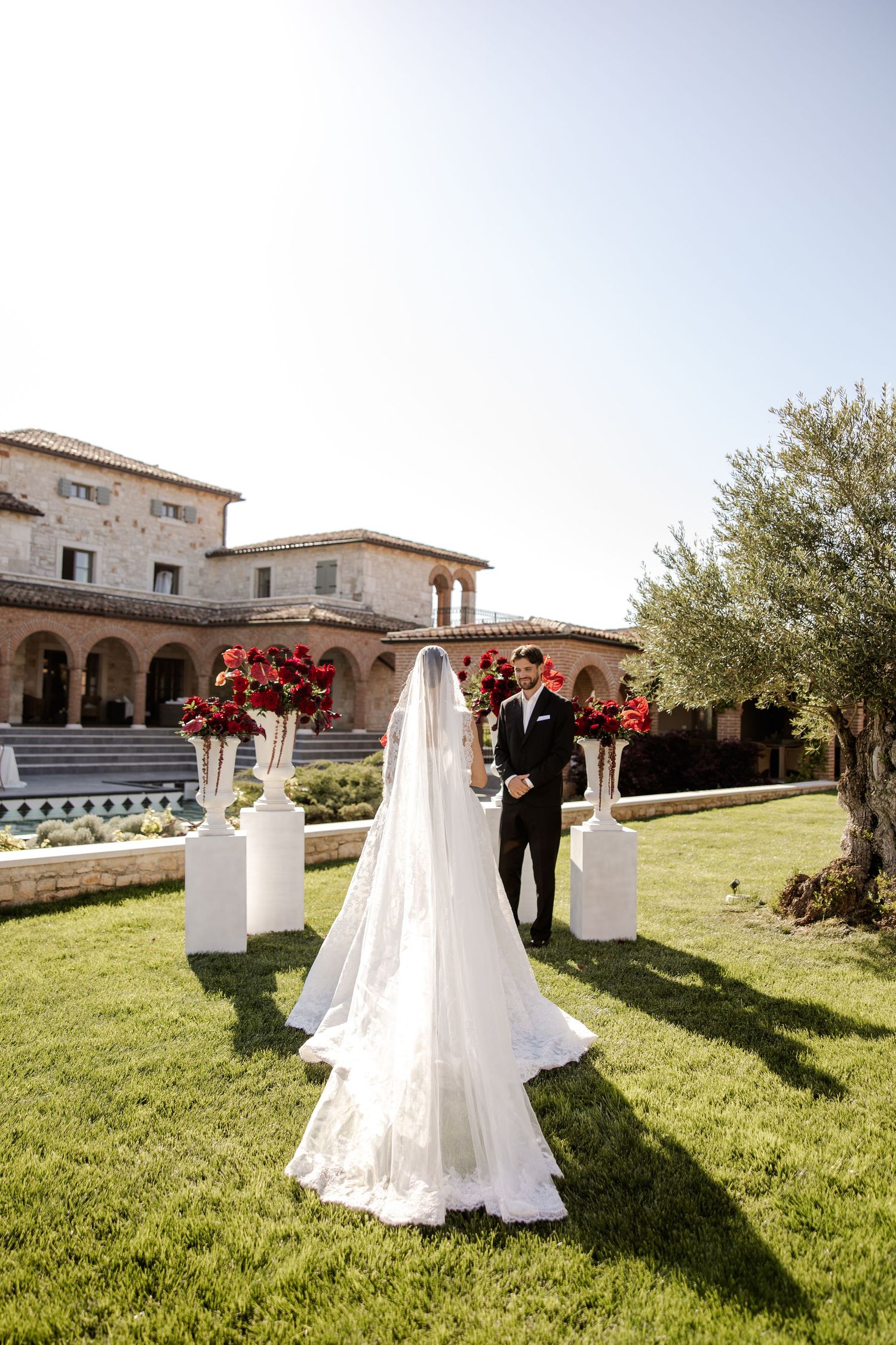 Bride in white gown and veil walks toward groom at outdoor wedding ceremony with red floral arrangements and stone villa.