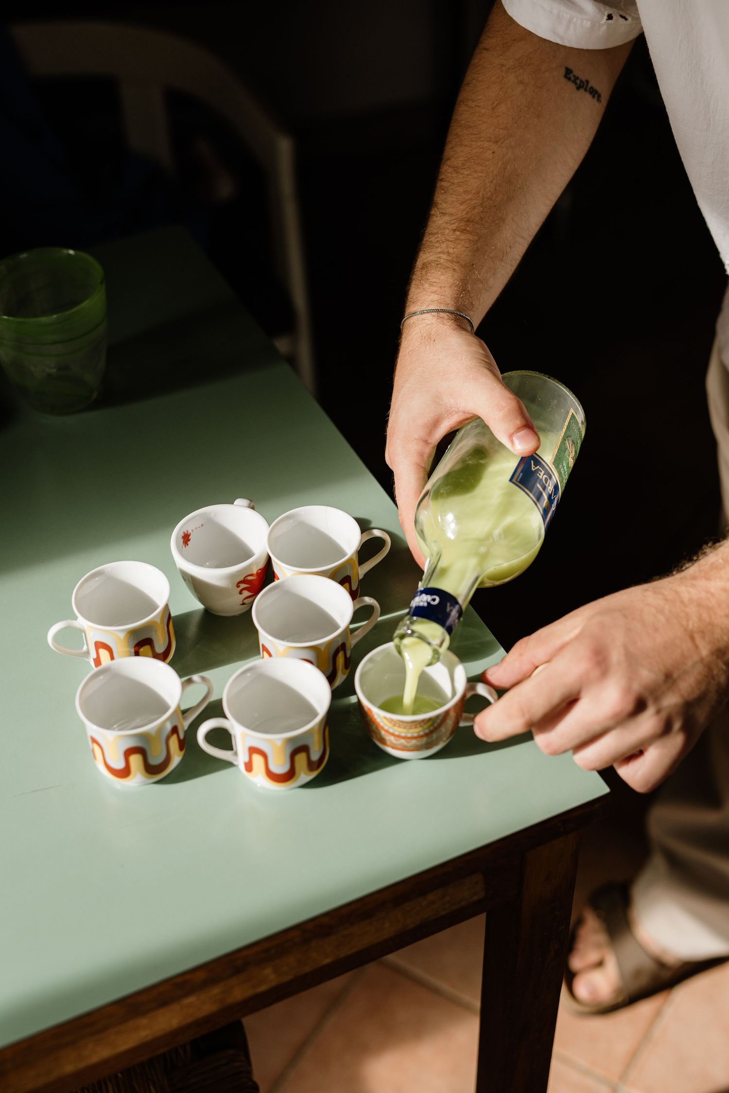 Man pouring green liquid from bottle into decorative ceramic mugs arranged on a light green table.