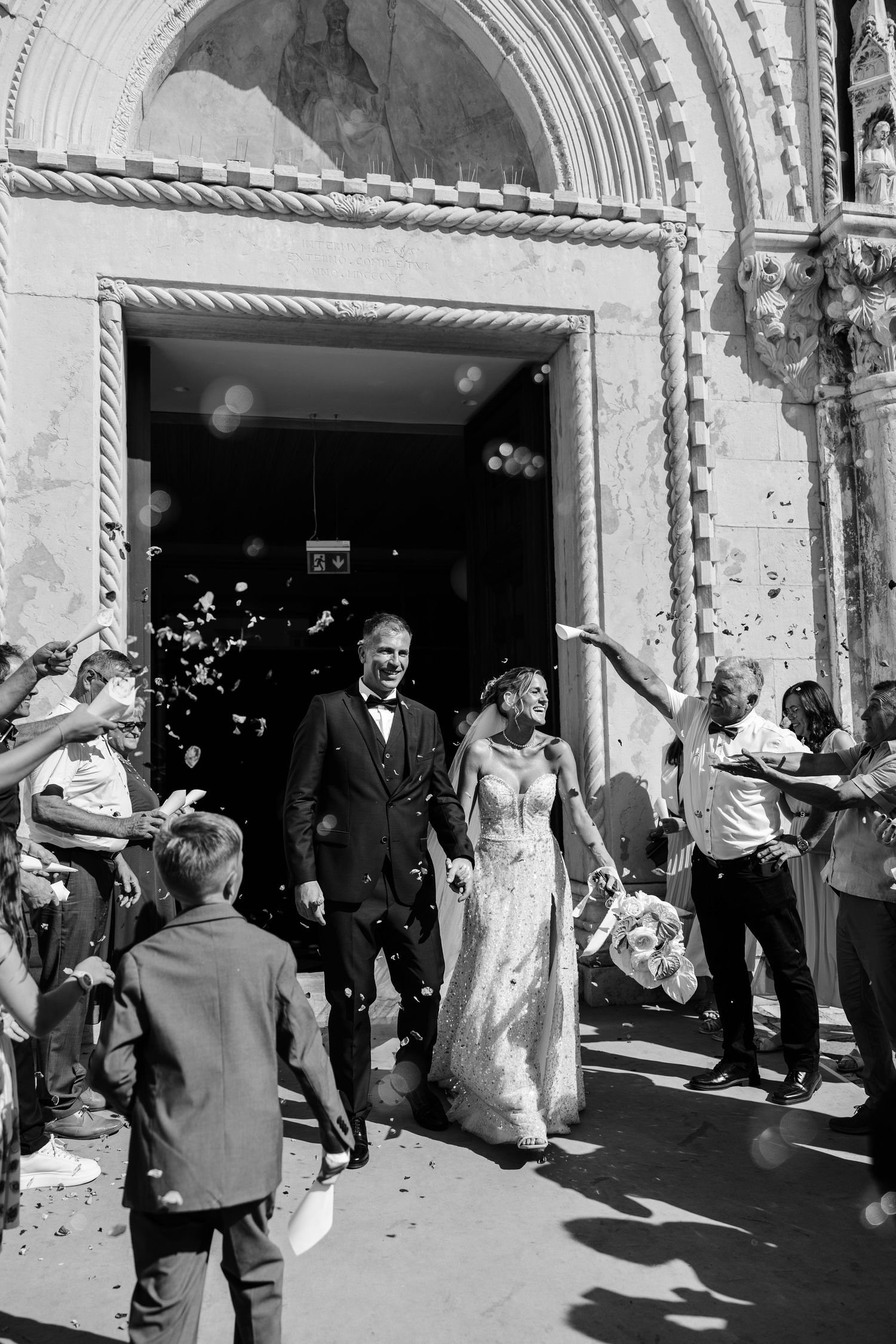 Black and white photo of newlyweds exiting church amid confetti shower, guests celebrating outside ornate stone doorway.
