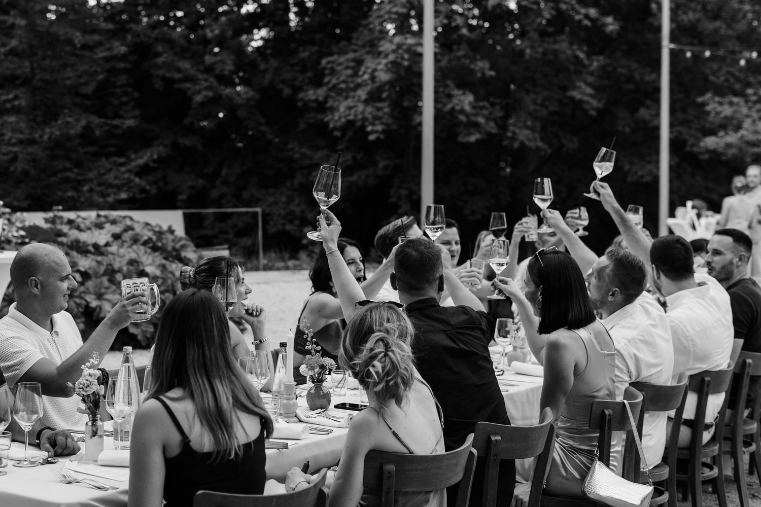 Guests raising wine glasses in a toast at an outdoor wedding reception dinner in black and white.