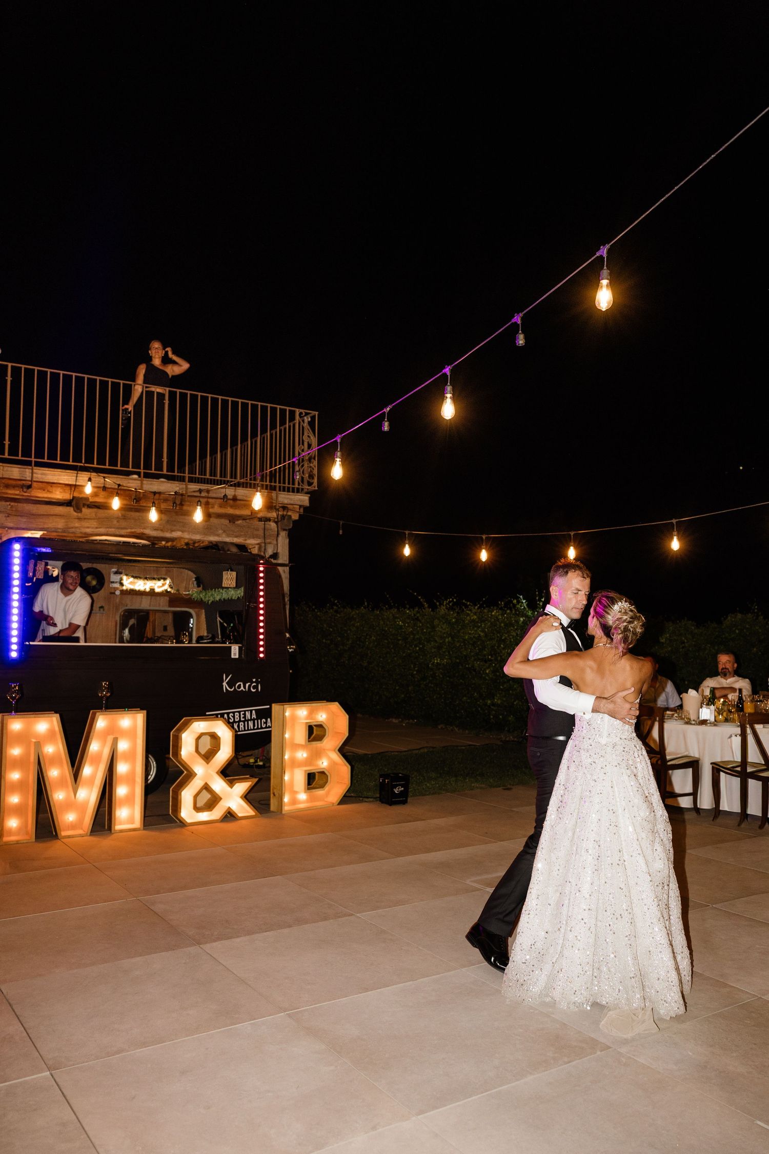 Newlyweds share first dance under string lights beside illuminated M&B marquee letters at outdoor night reception.