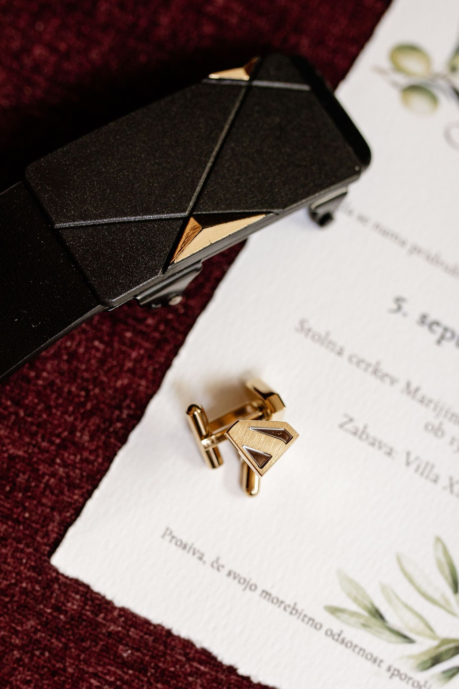 Gold cufflinks and monogram initial beside a black belt and wedding invitation on burgundy fabric.