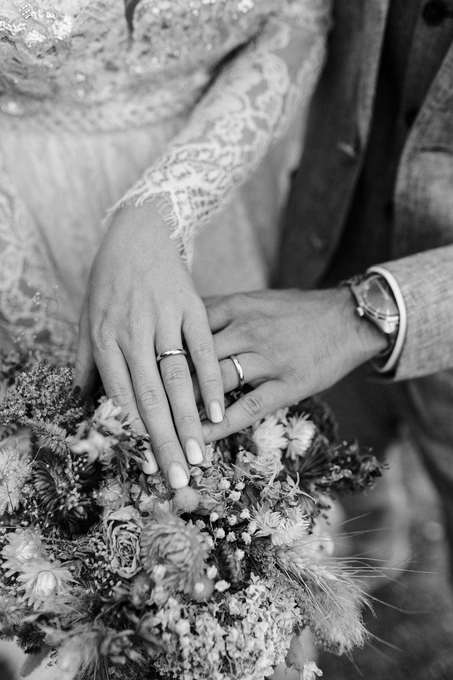 Black and white close-up of newlyweds hands with wedding rings resting on a rustic bridal bouquet.