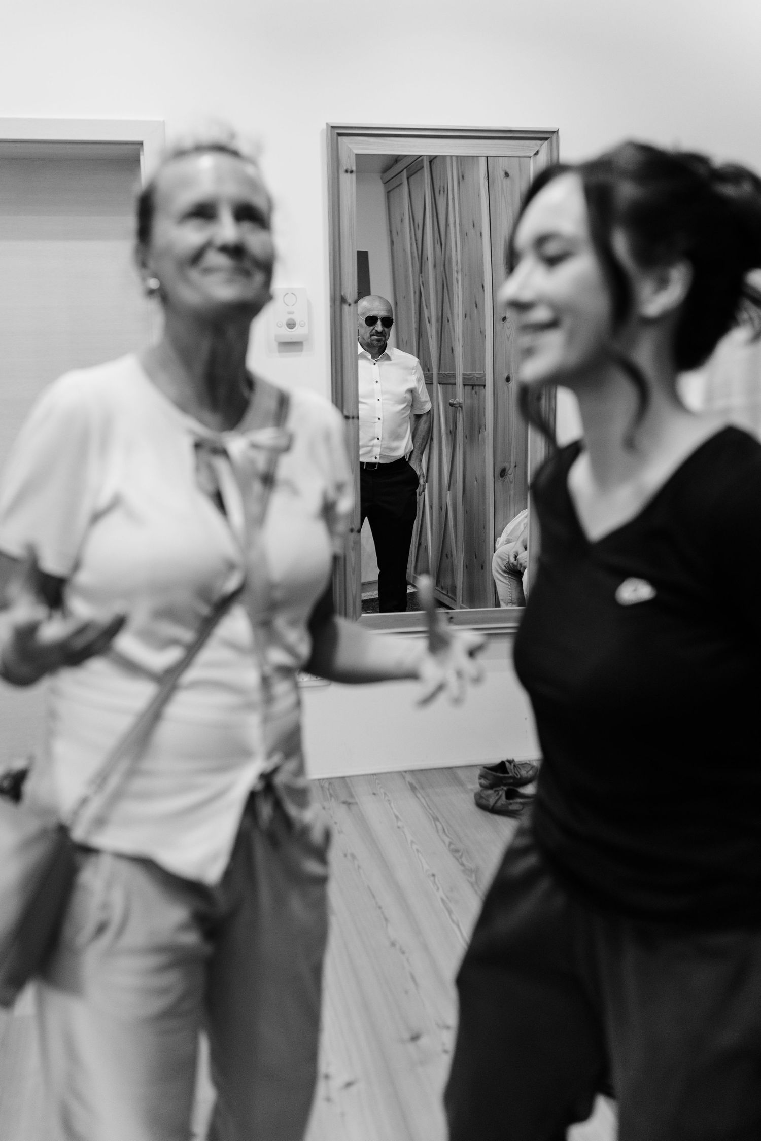 Black and white photo of two women smiling indoors while a man in sunglasses watches from a doorway.