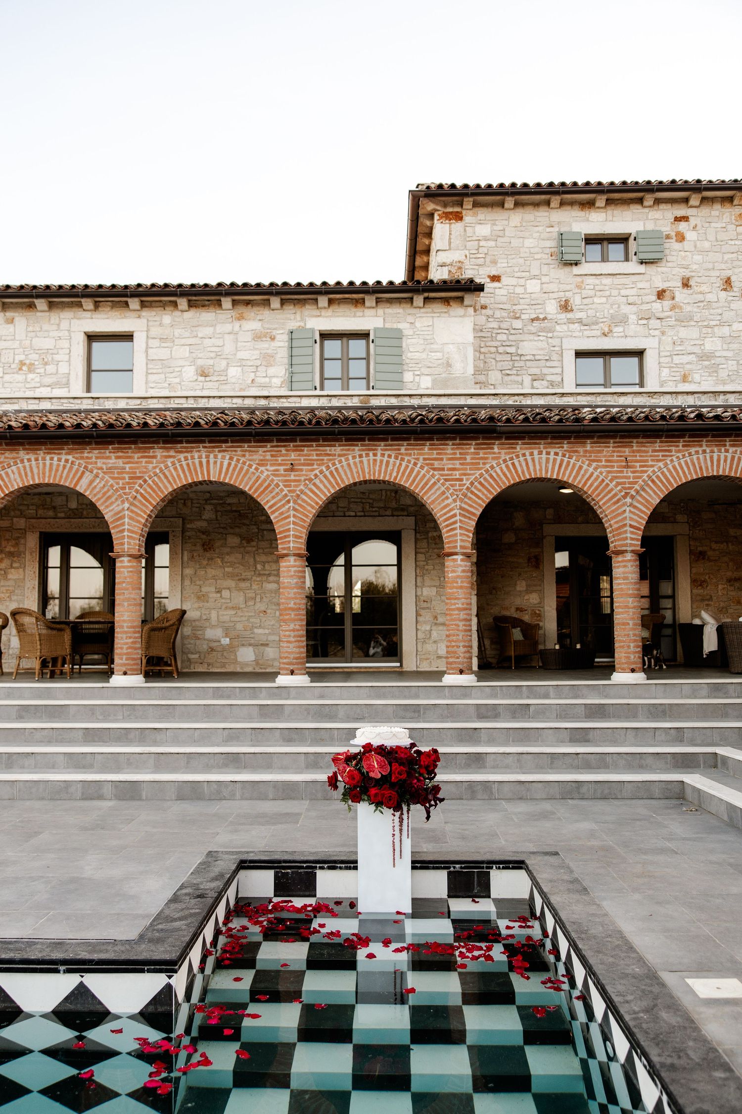 Elegant Italian villa courtyard with brick arches, checkered fountain, and red rose centerpiece arrangement.