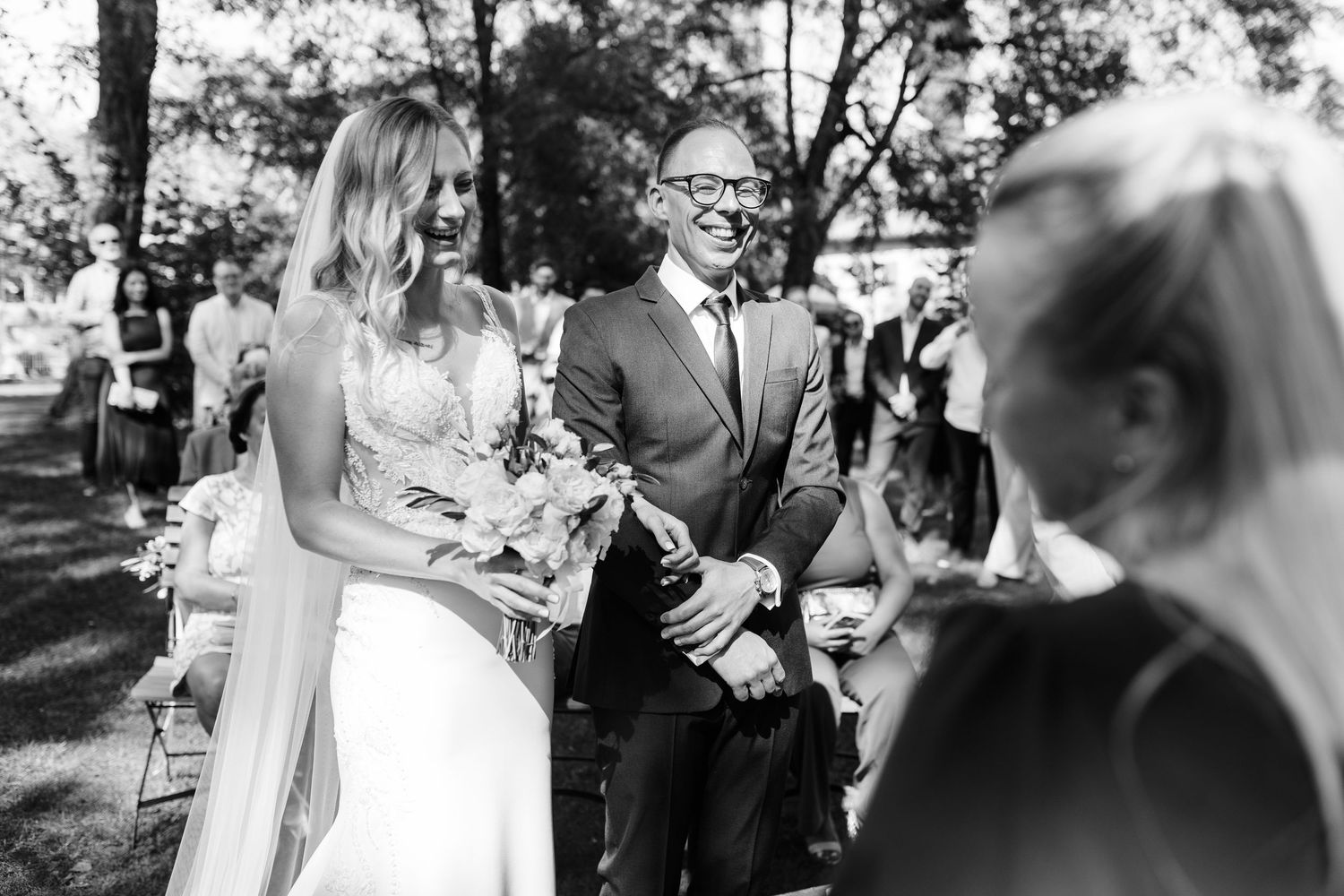 Black and white photo of a smiling bride walking down the aisle with an escort at an outdoor wedding ceremony.
