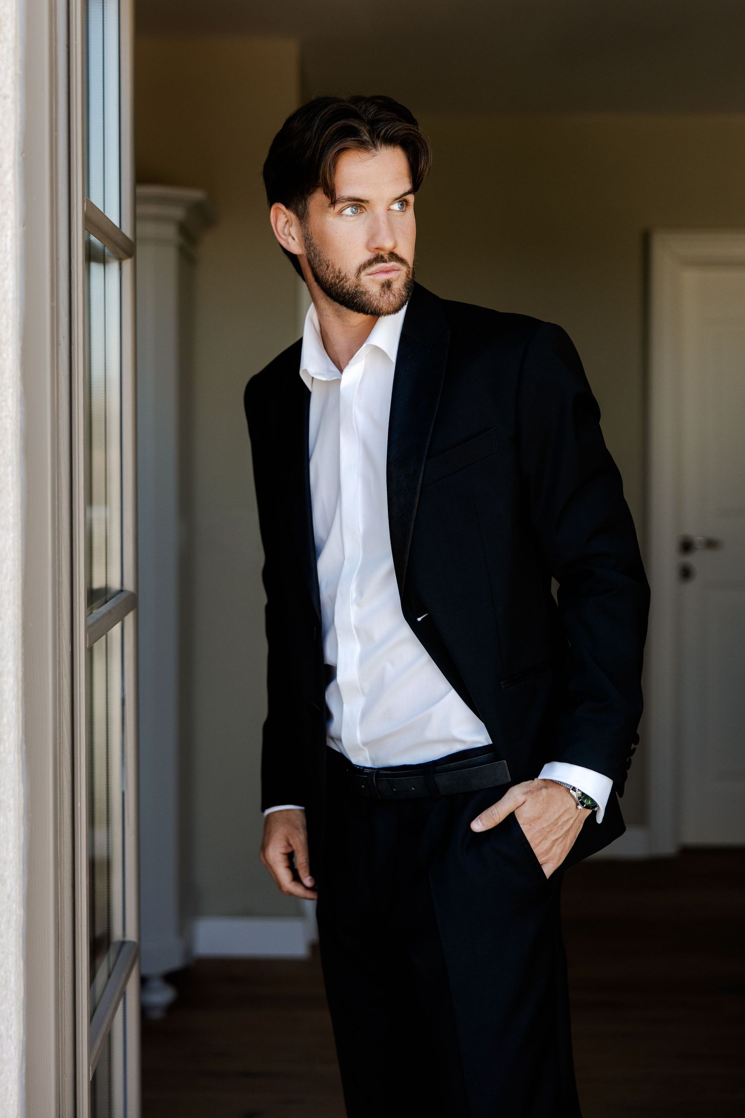 A well-dressed man in a black suit and white shirt stands by a window, looking away thoughtfully.