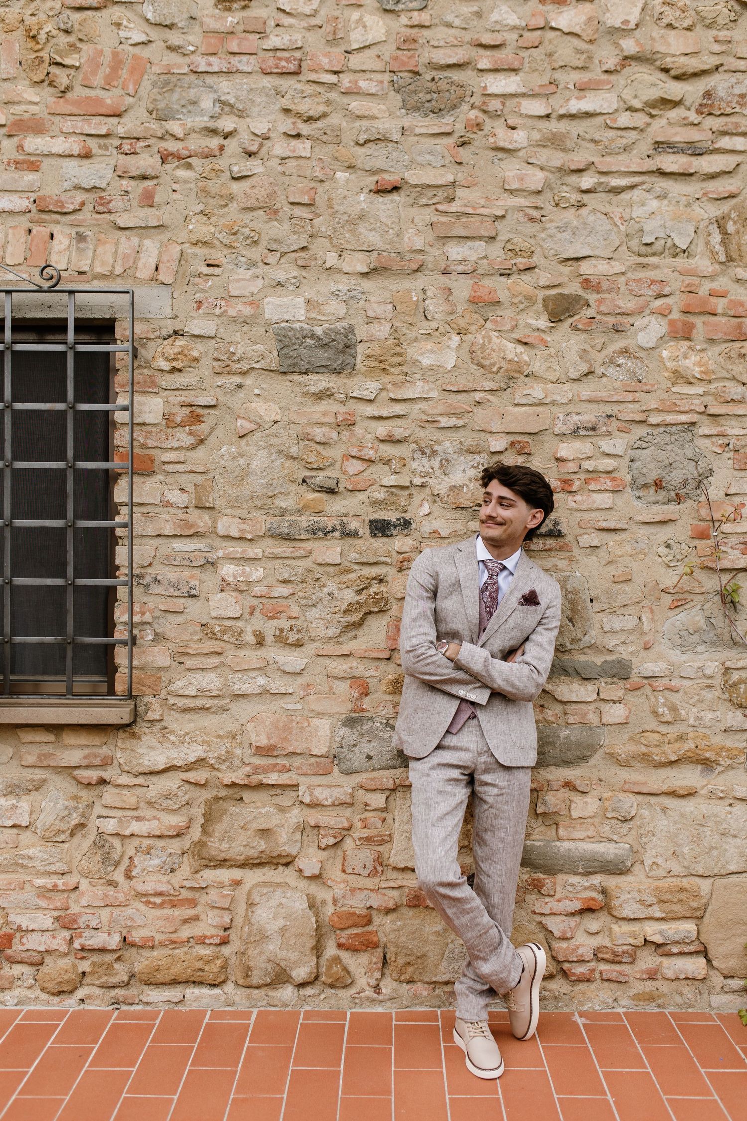 Smiling groom in a beige linen suit leaning against a rustic stone wall in Tuscany, Italy.