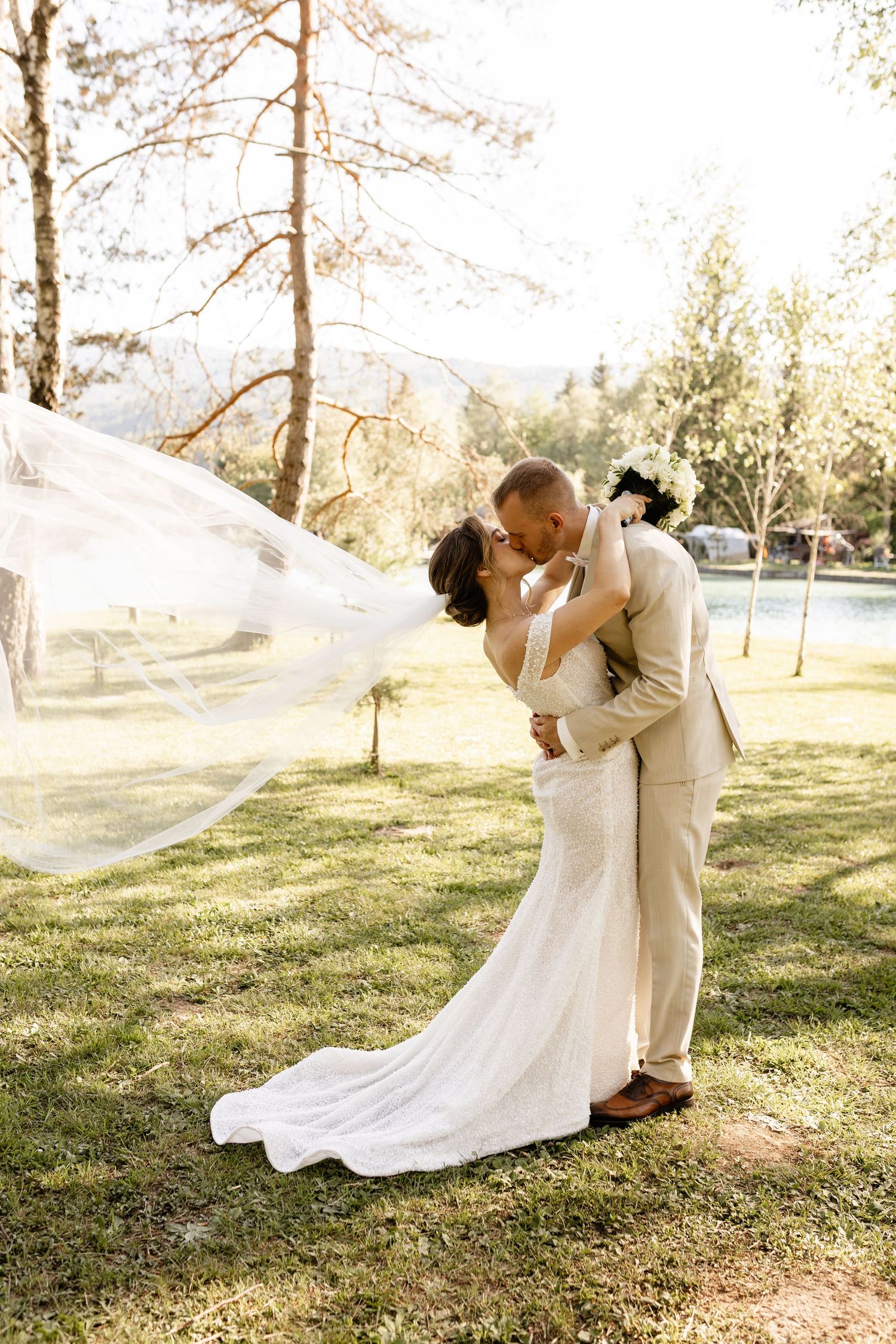 Bride and groom kiss outdoors with flowing veil caught in wind against sunlit trees and green grass.