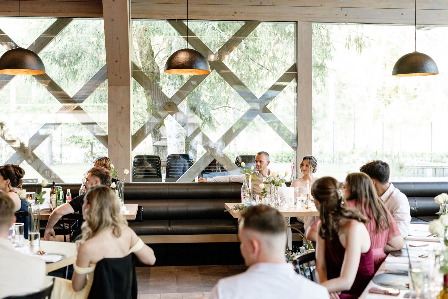 Guests gathered at elegant restaurant tables during a wedding reception with pendant lights and large windows.