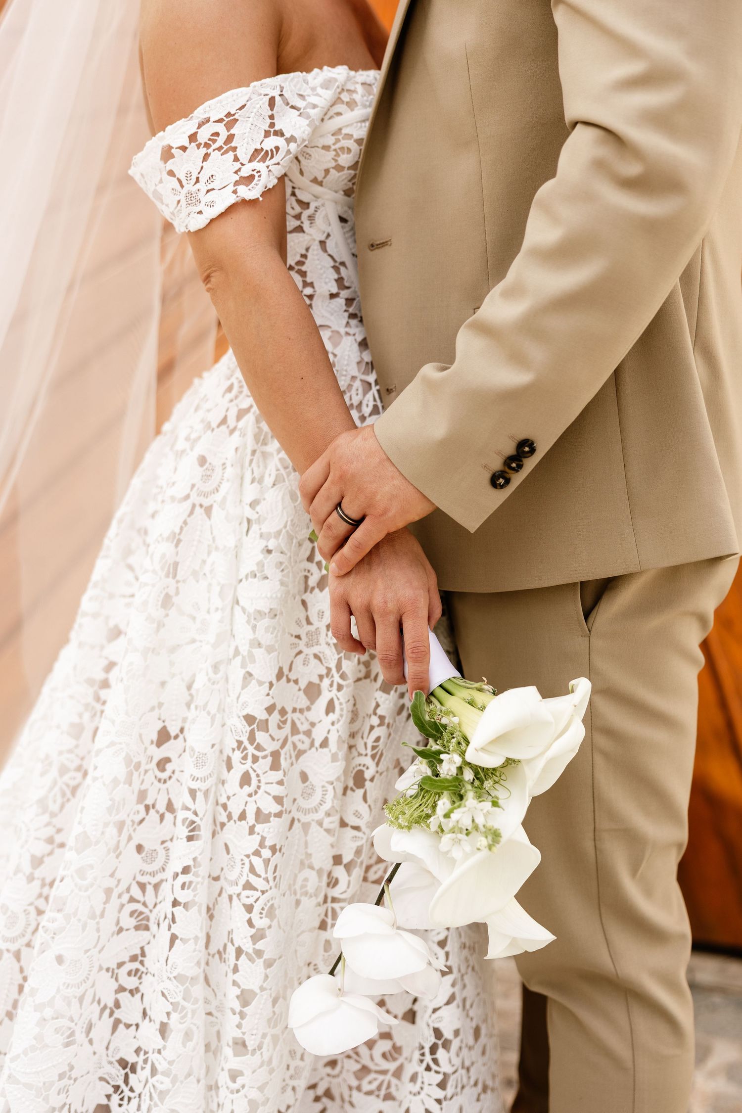 Bride in lace off-shoulder gown holds groom's hand and white calla lily bouquet, both in elegant wedding attire.