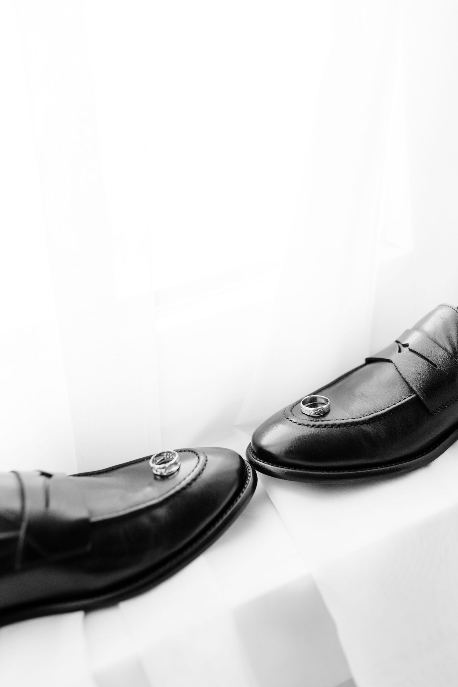 Black leather loafers with silver wedding rings resting on them, displayed on a white surface in black and white.