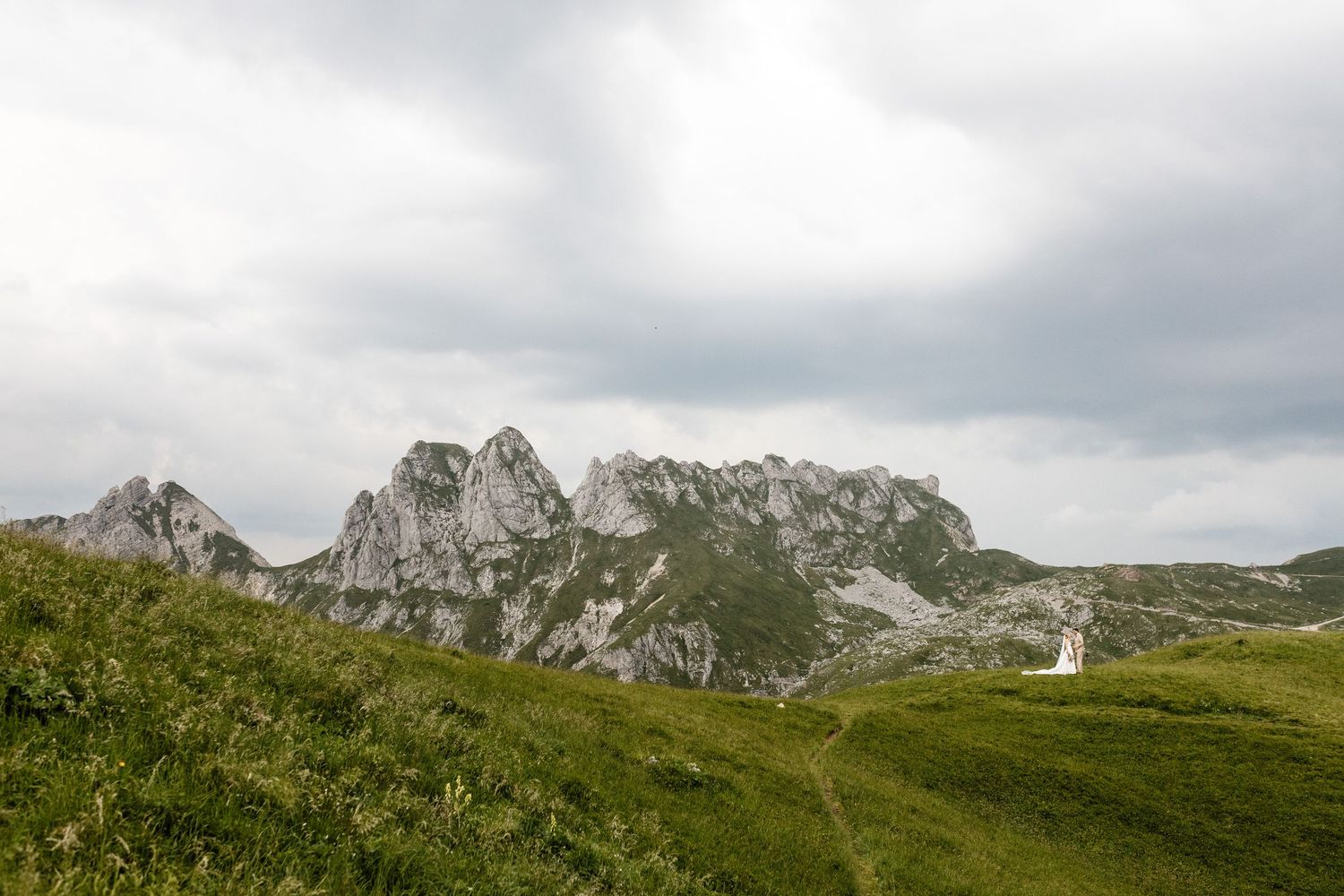 Bride and groom appear as tiny figures on vast green Alpine meadow with dramatic rocky mountain peaks behind them.