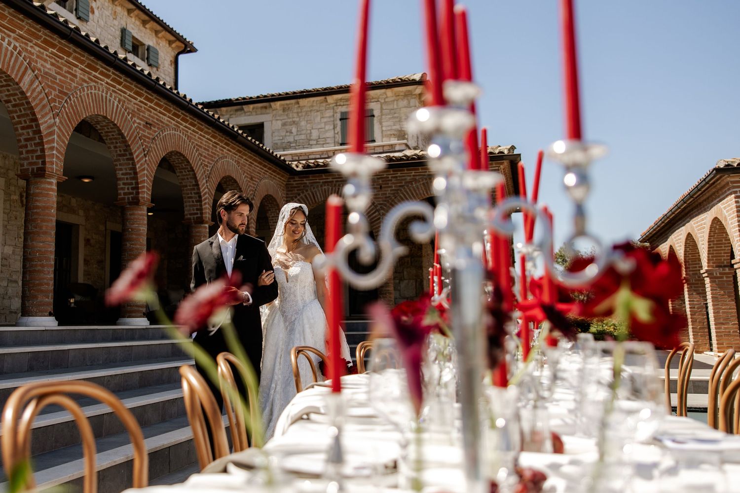 Elegant outdoor wedding reception with red candelabras, a couple descending stone steps at a rustic Italian villa.