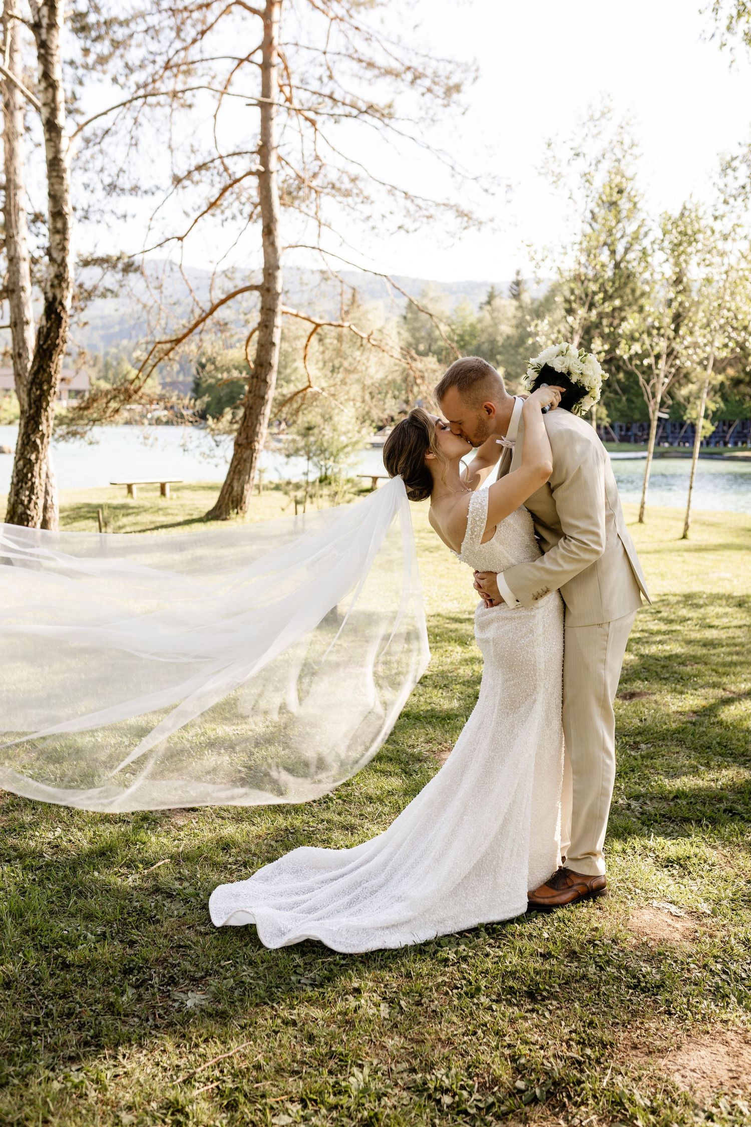 Bride and groom share a romantic kiss outdoors, with flowing veil, white dress, and beige suit near a lake.