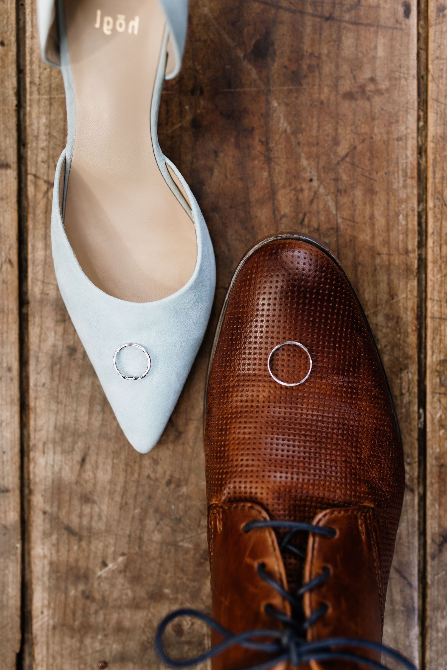 Wedding rings resting on light blue heels and brown leather oxford shoes on rustic wooden floor.